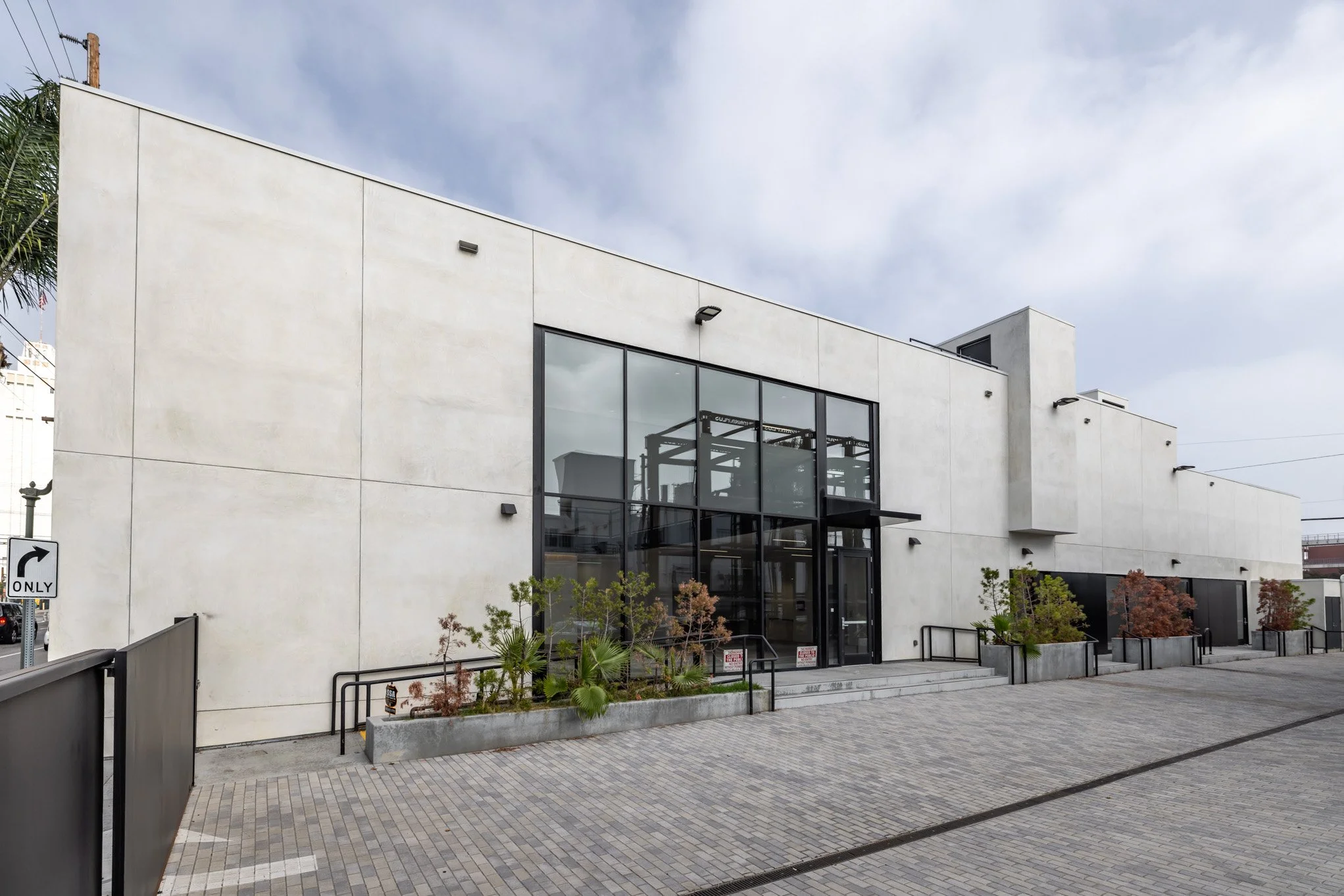 Modern commercial building with large glass entrance and minimalistic design, surrounded by small landscaped planters and brick pavement.