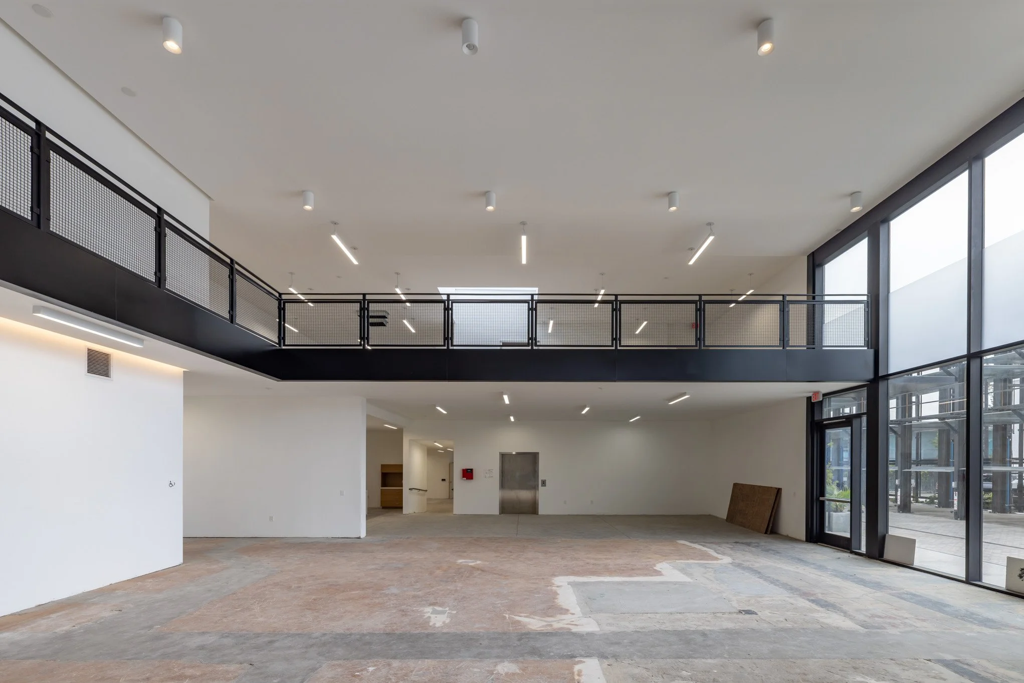 Interior view of an empty commercial space with large floor-to-ceiling windows, black railings on the second level, and unfinished flooring.