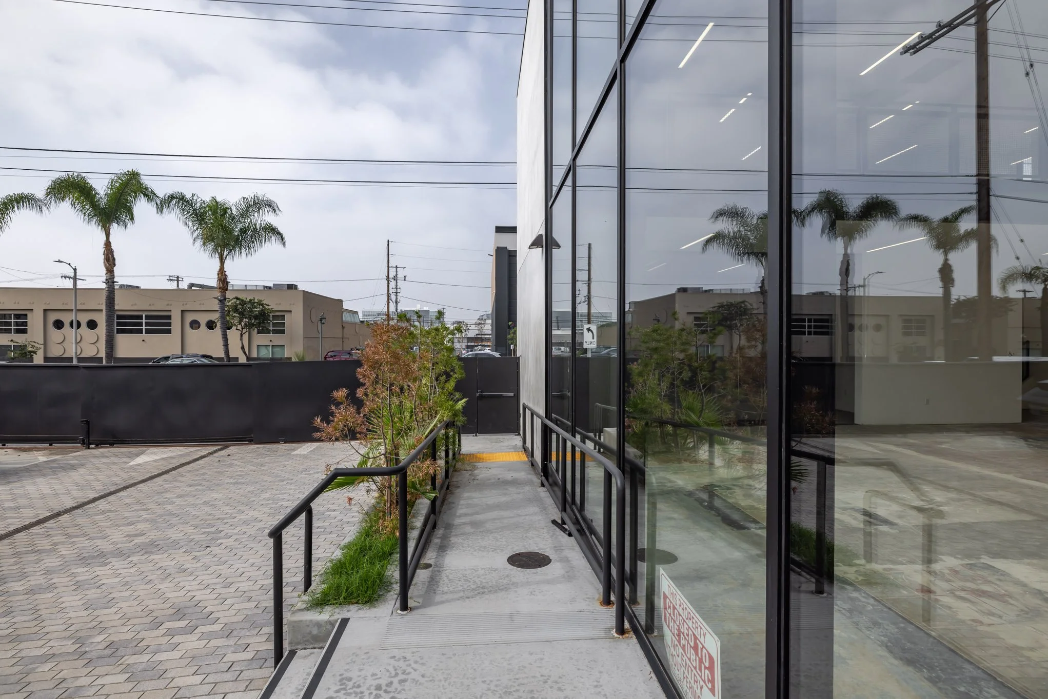 Sidewalk with black metal handrails in front of a glass building, with a parking lot and costumed buildings with palm trees in the background.