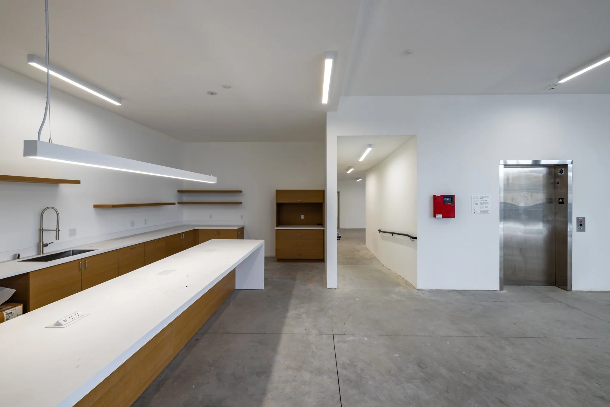 Empty modern kitchen with white countertops and wooden cabinets, stainless steel sink, shelving, and an elevator in the background.