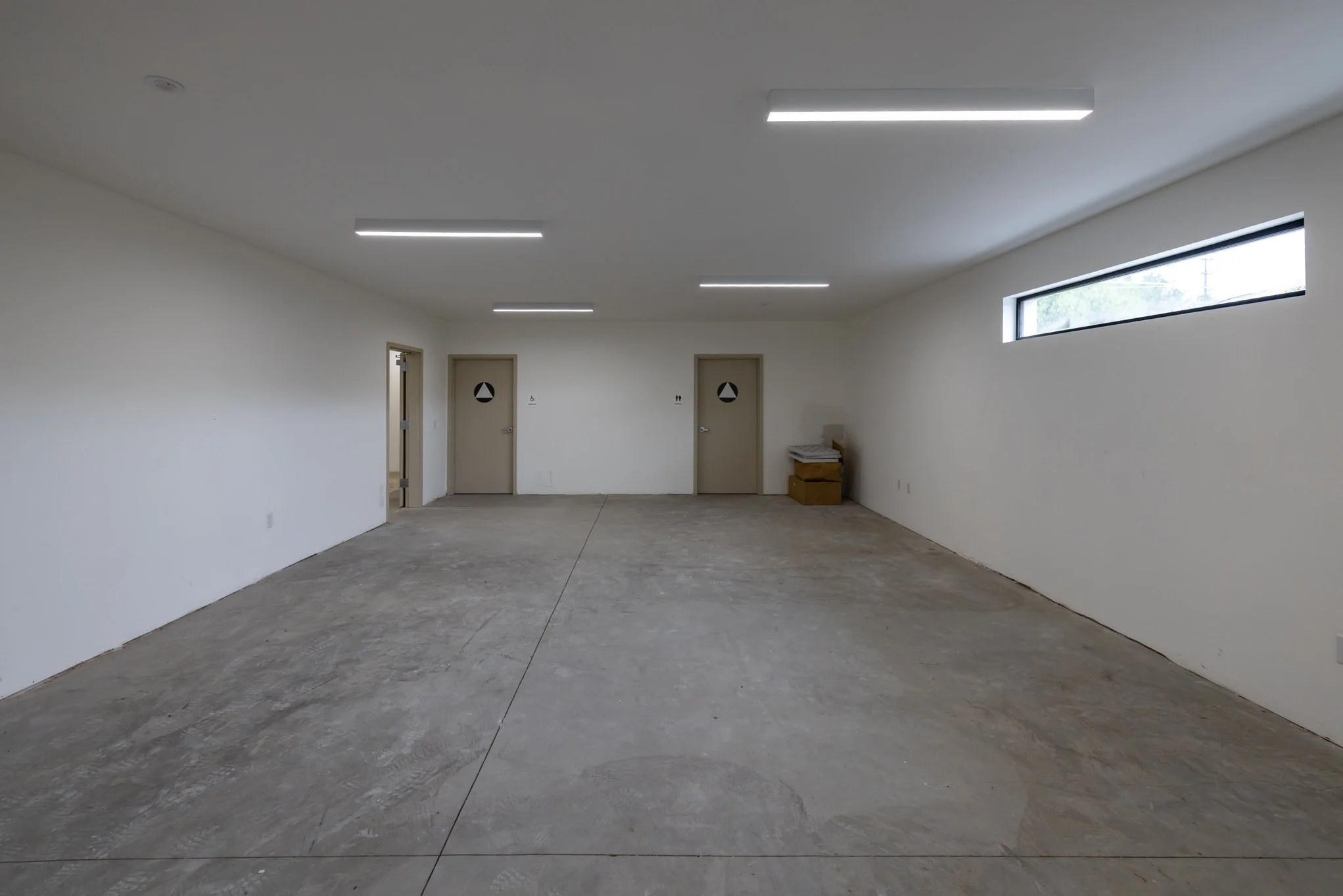 Empty room with concrete floor, white walls, fluorescent ceiling lights, and small horizontal window, with two labeled bathroom doors at the back.
