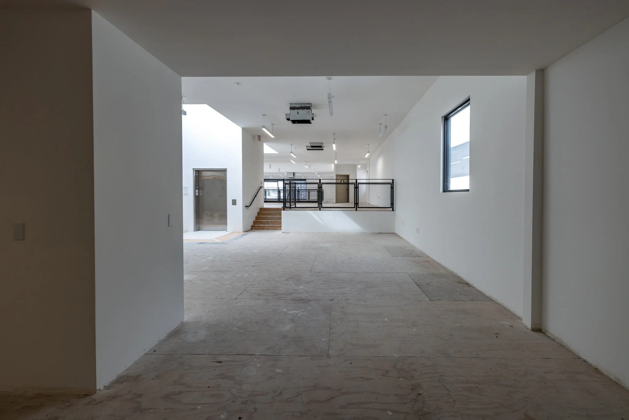 Empty interior space with white walls and unfinished flooring, a window on the right wall, stairs leading up, and an elevator door on the left.