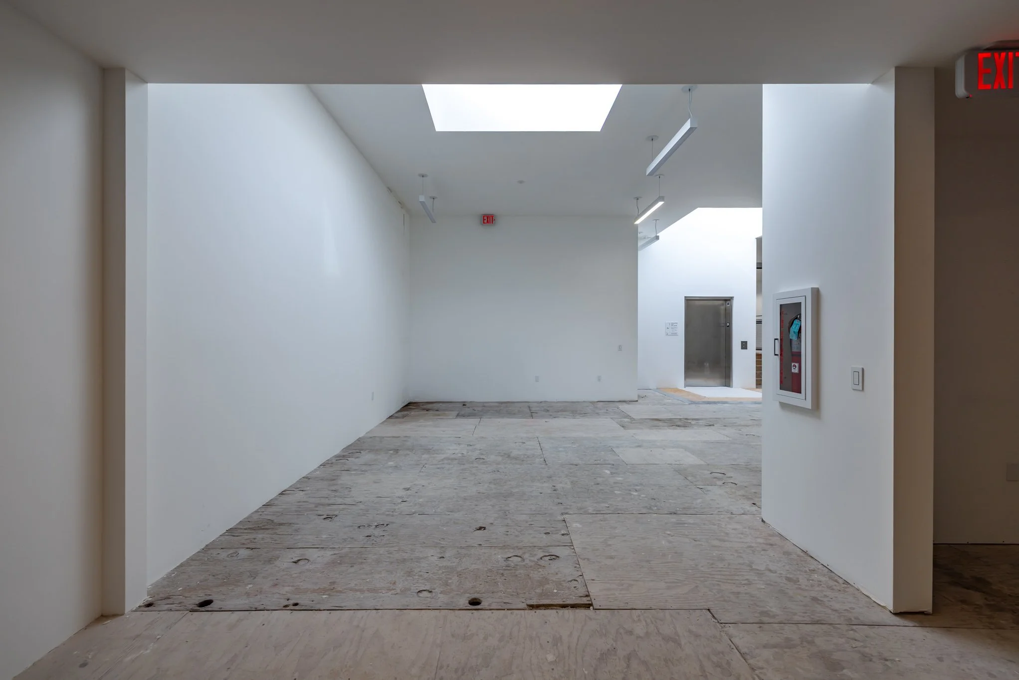 Empty interior space with white walls and exposed flooring, elevator in the background, and emergency exit signs.