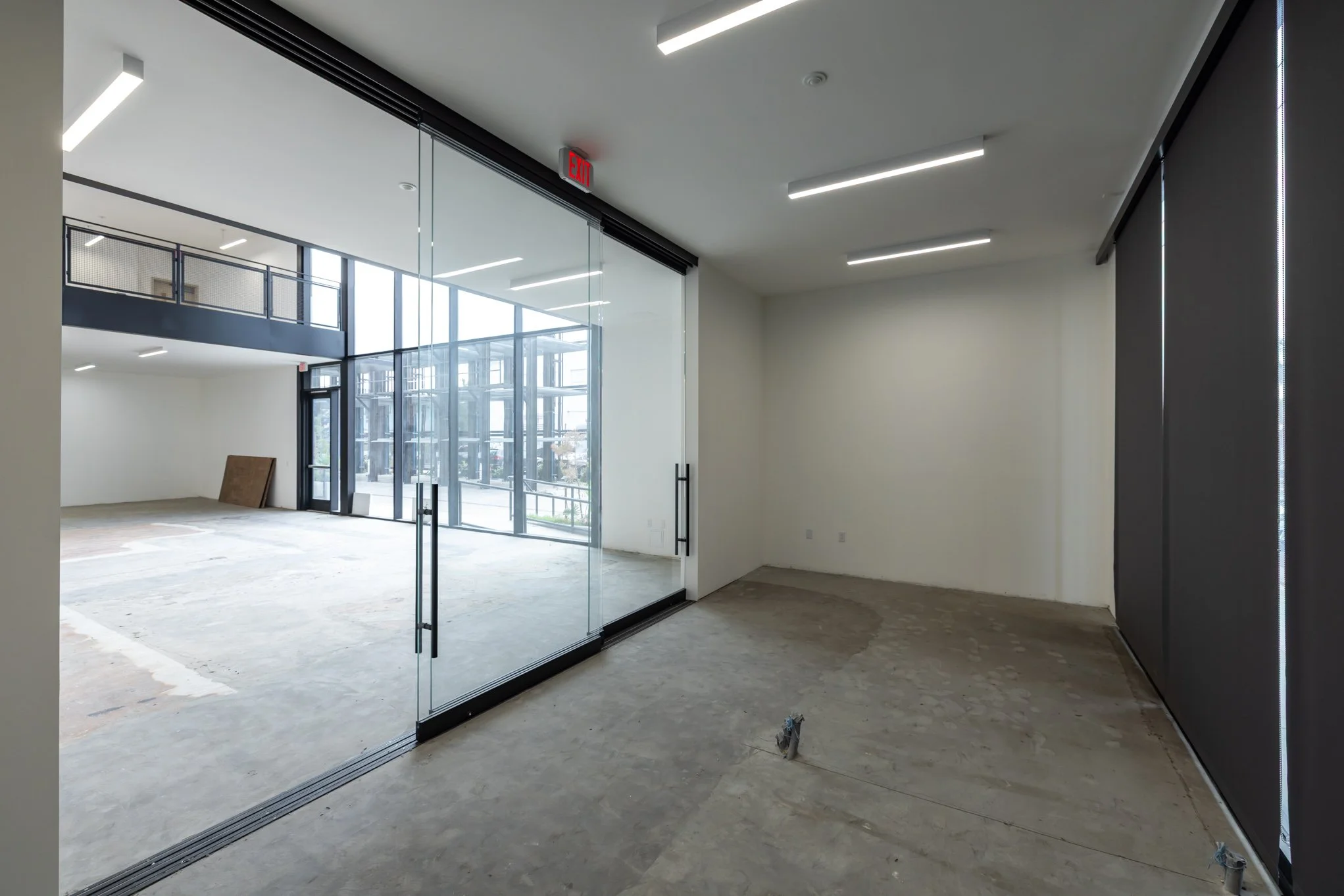 Empty commercial space with large glass entrance doors, modern ceiling lights, and an upstairs balcony visible through the glass. The floor is unfinished, and there's a black wall with vertical LED strip lights on the right.