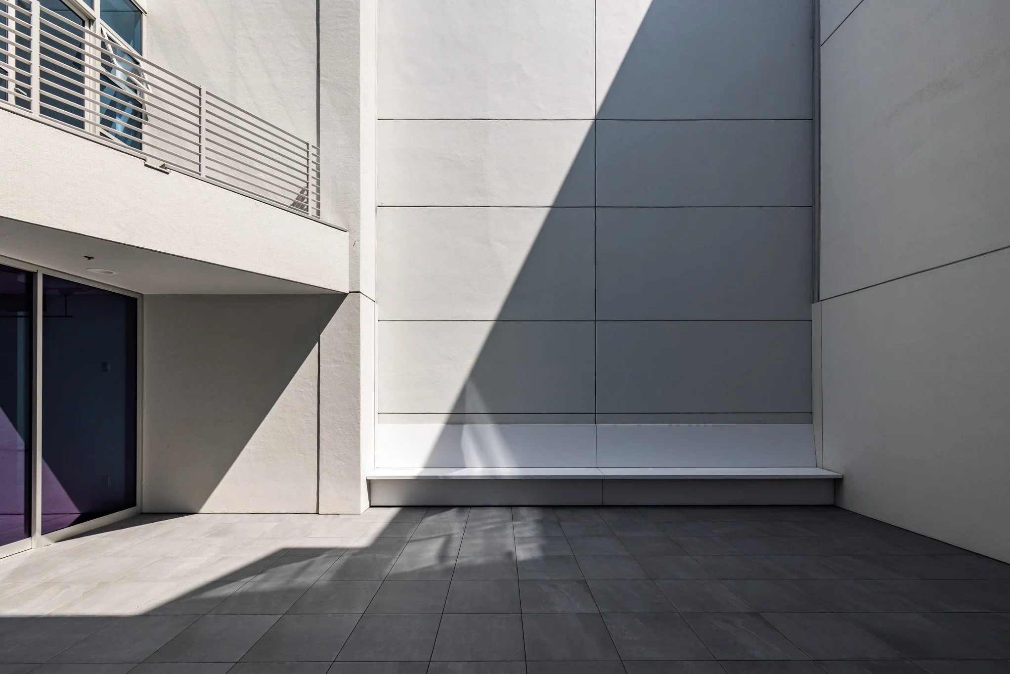 Modern minimalist courtyard with white walls, a glass door, and a balcony with metal railings, partially shaded by a large shadow.