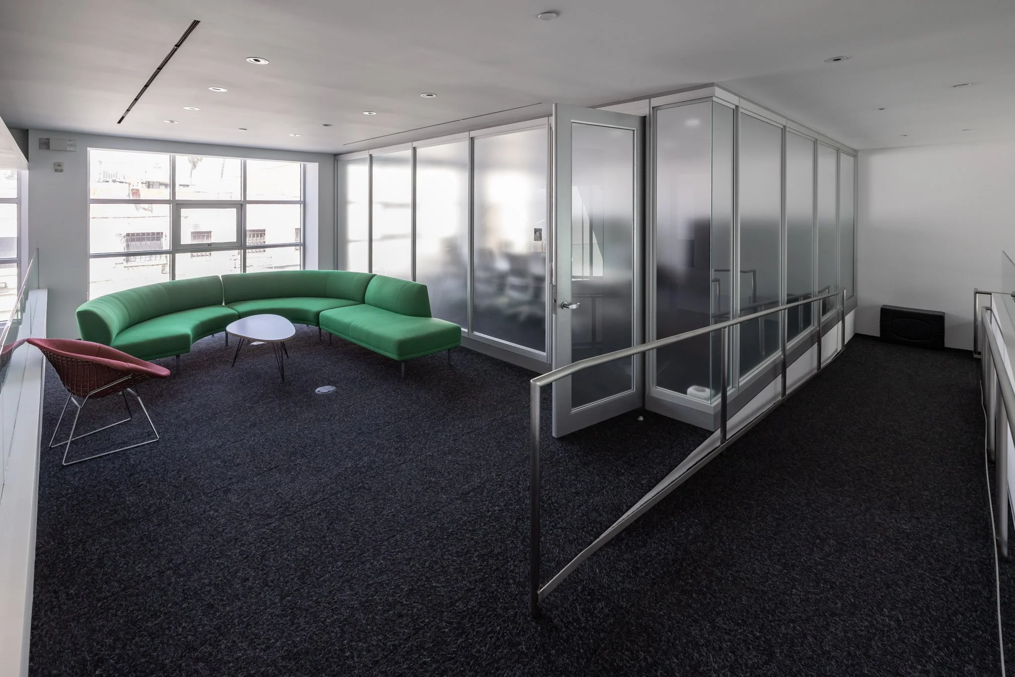 Modern office lounge with a green semi-circular sofa, a pink chair, and a small round coffee table, next to glass partitions and large windows.