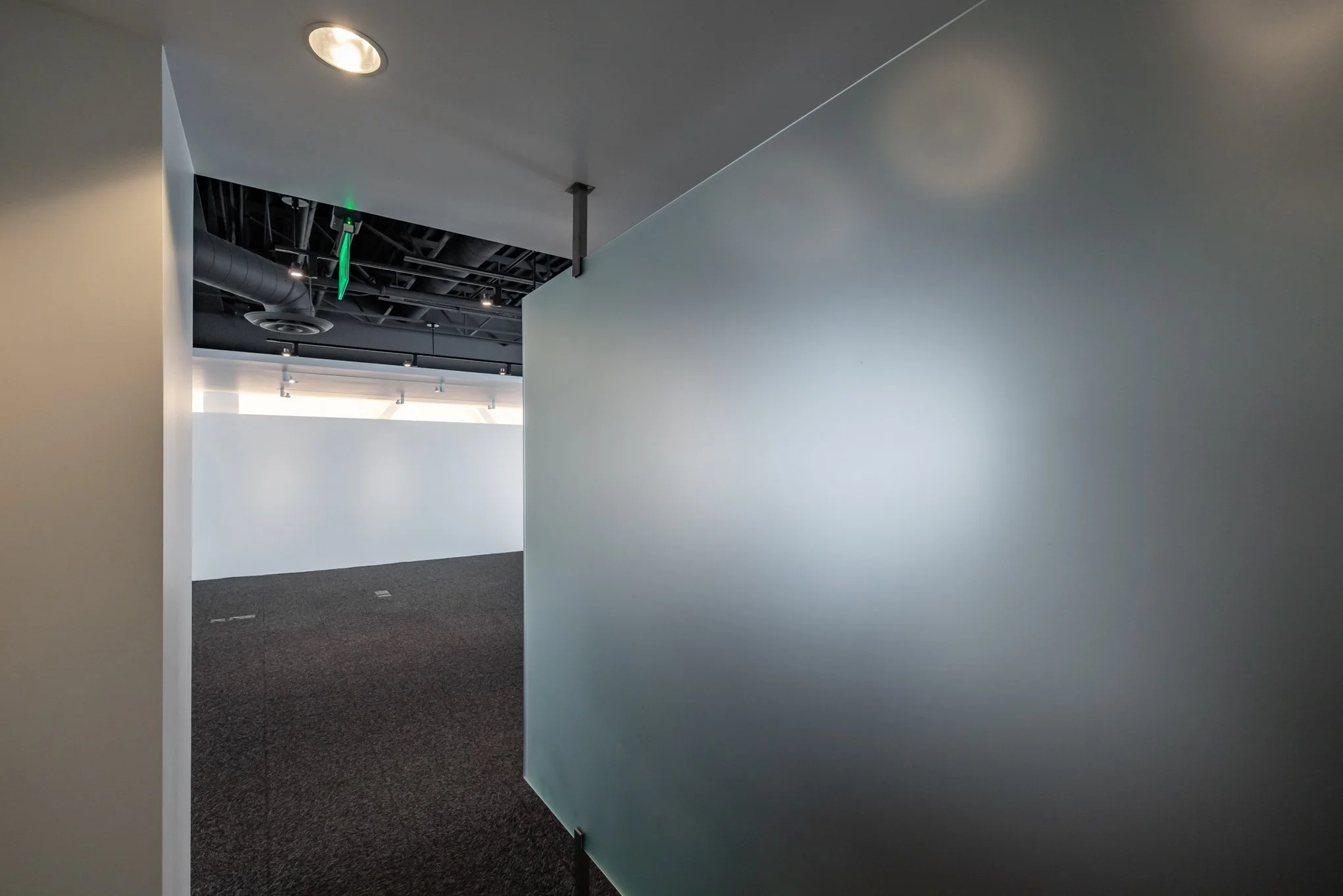 Empty, modern office corridor with gray wall on the right, dark carpeted floor, white wall visible at the end, ceiling with lighting and HVAC vents, and a green emergency exit sign hanging from the ceiling.