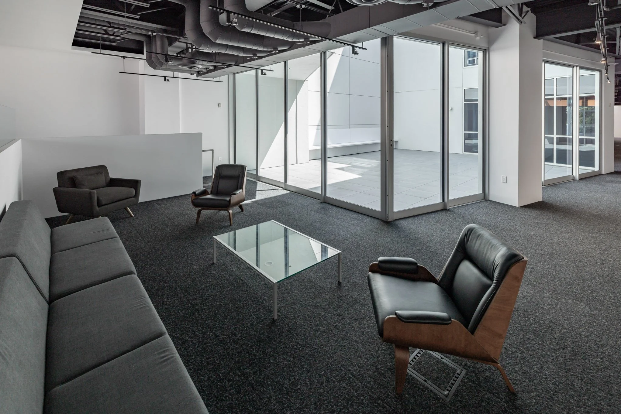 Modern office lounge area with black and gray chairs, a gray sofa, a glass coffee table, and large windows with sliding doors leading to an outdoor patio.