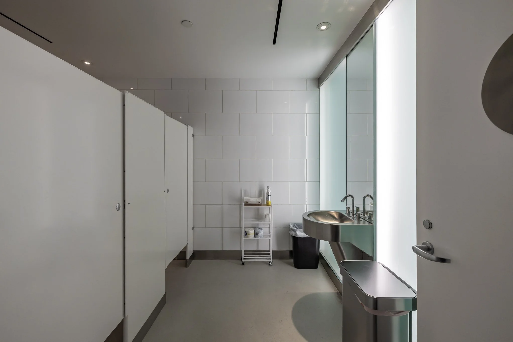A clean, minimalist public restroom with white tile walls, a stainless steel sink, a trash can, a small rolling cart with supplies, and a frosted glass window.