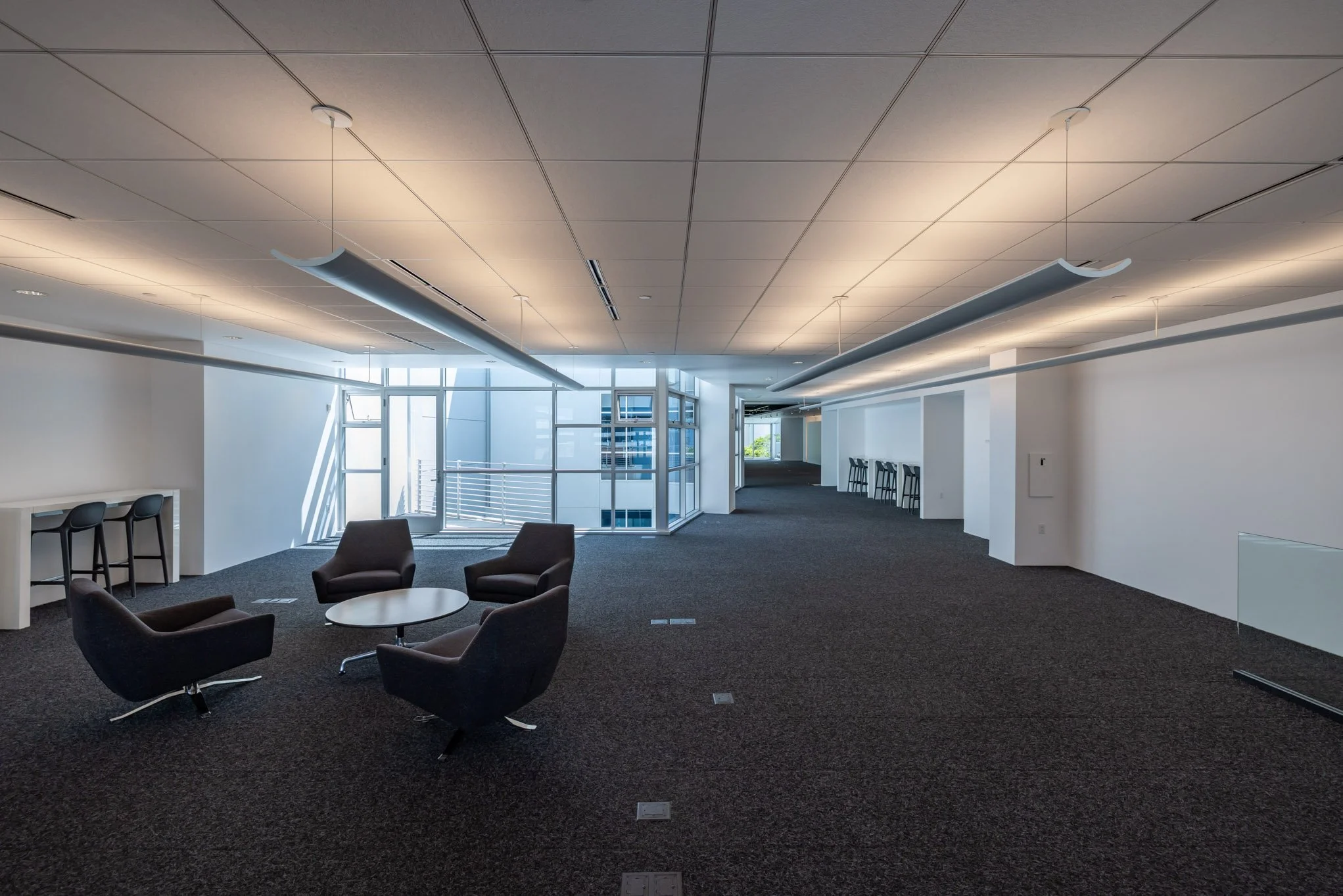 Empty modern office space with black chairs around a small round table, tall windows, and a long corridor with barstools.