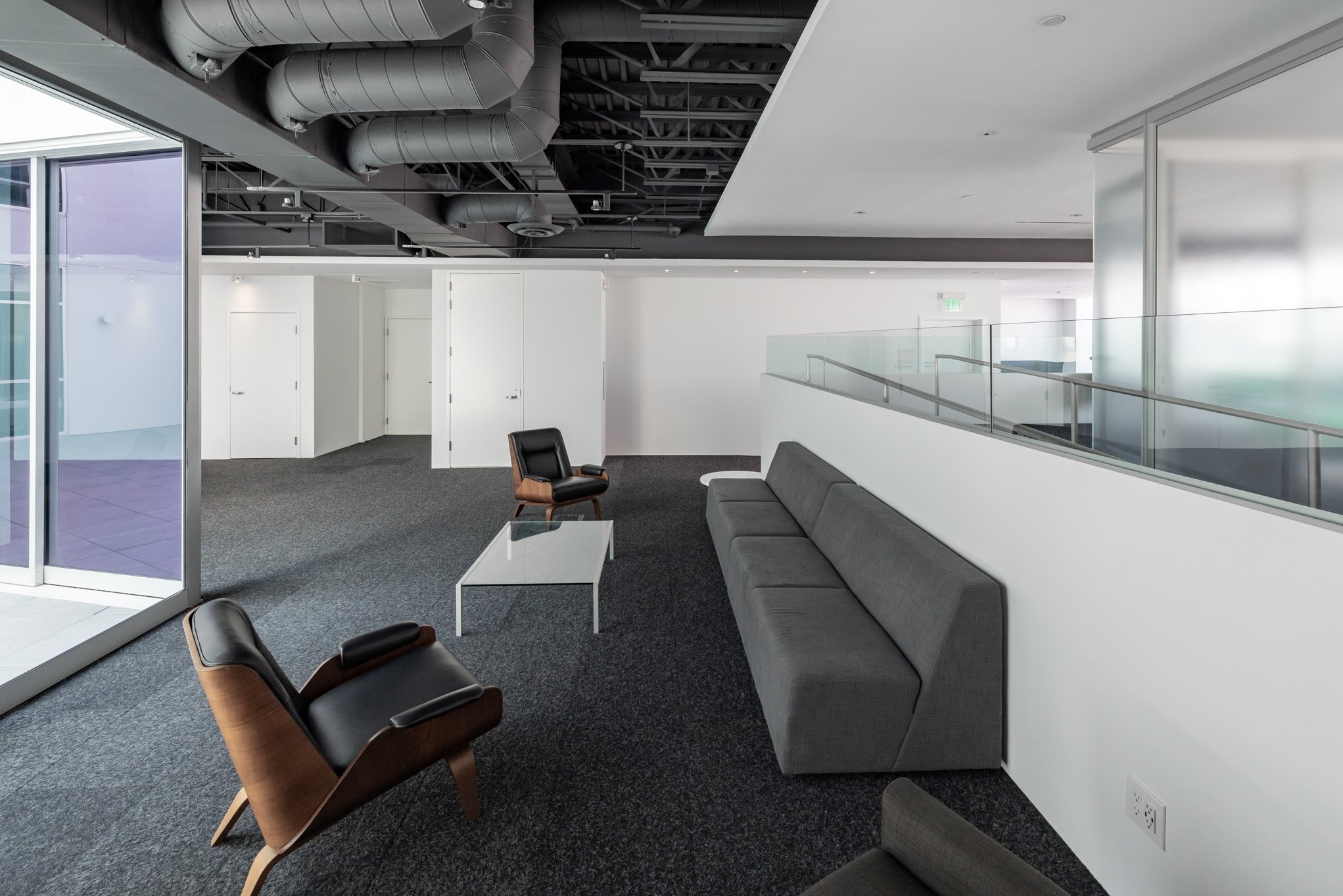 Modern office lounge area with black, gray, and brown chairs, a white coffee table, white walls, and glass railing, illuminated by natural light.