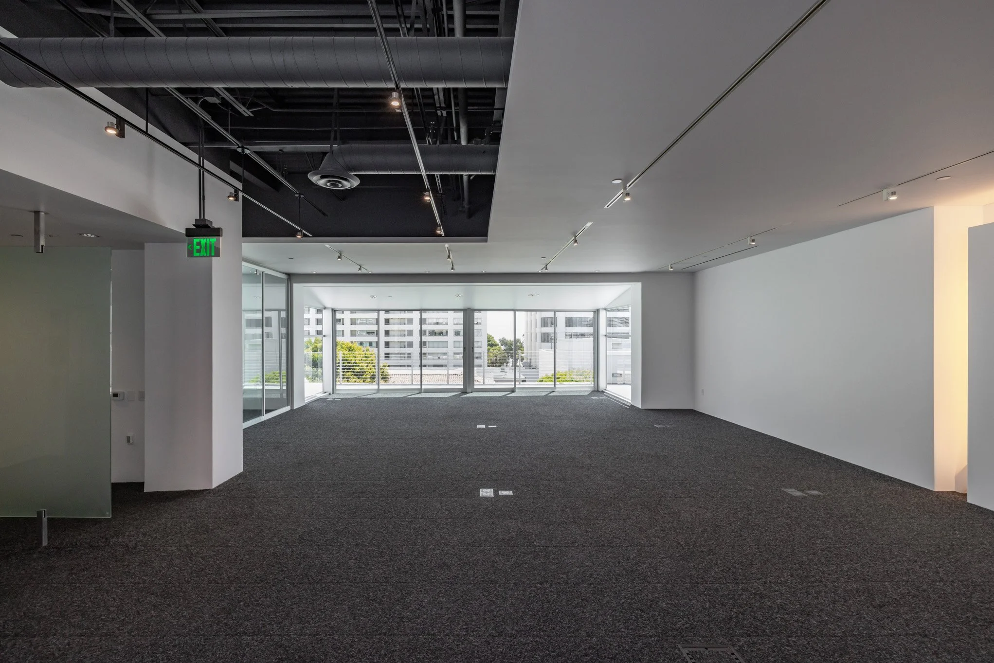 Empty office space with large windows overlooking city buildings, dark carpeted floor, white walls, and exposed ceiling with ventilation ducts and track lighting.