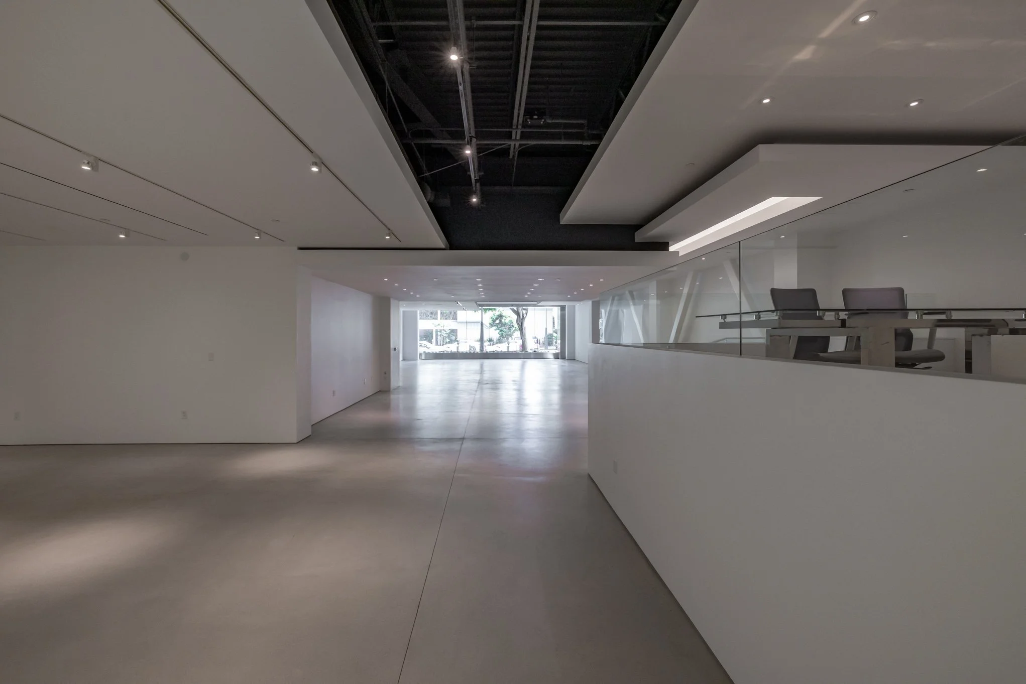 Empty modern interior space with white walls, polished concrete floor, and a reception or workspace area with chairs and glass panels, illuminated by ceiling lights.