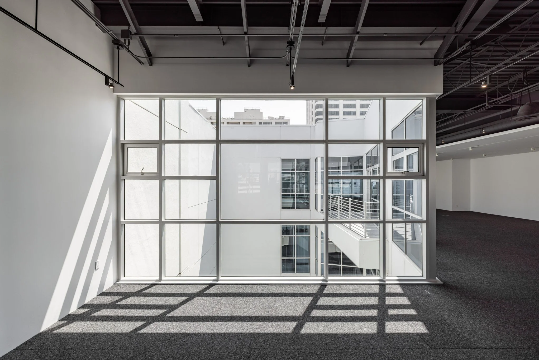 Empty modern office space with large floor-to-ceiling windows, casting shadows on the dark carpeted floor, and ceiling with exposed pipes and beams.