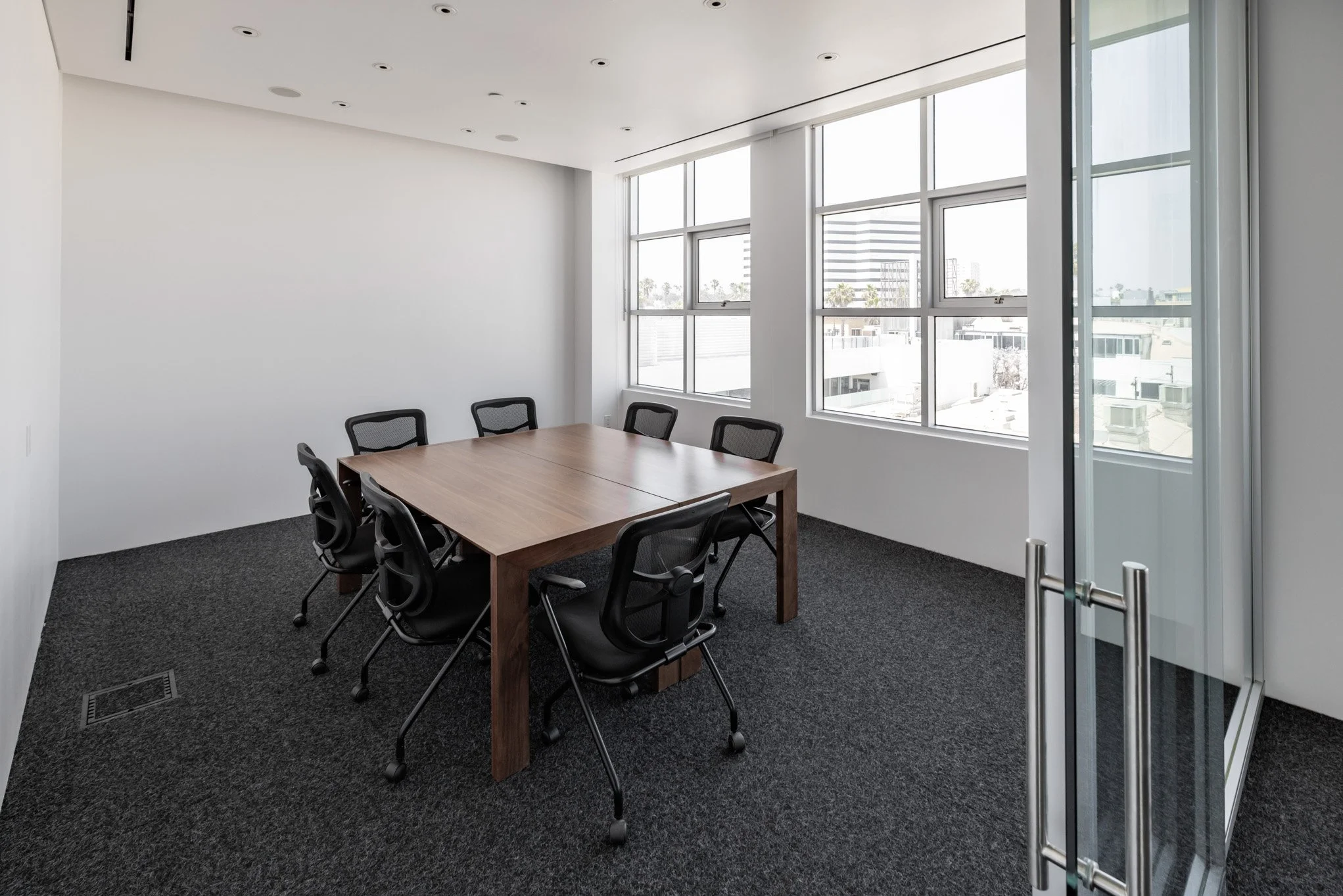 Empty office conference room with a wooden table and six black chairs, large windows, and a sliding glass door.