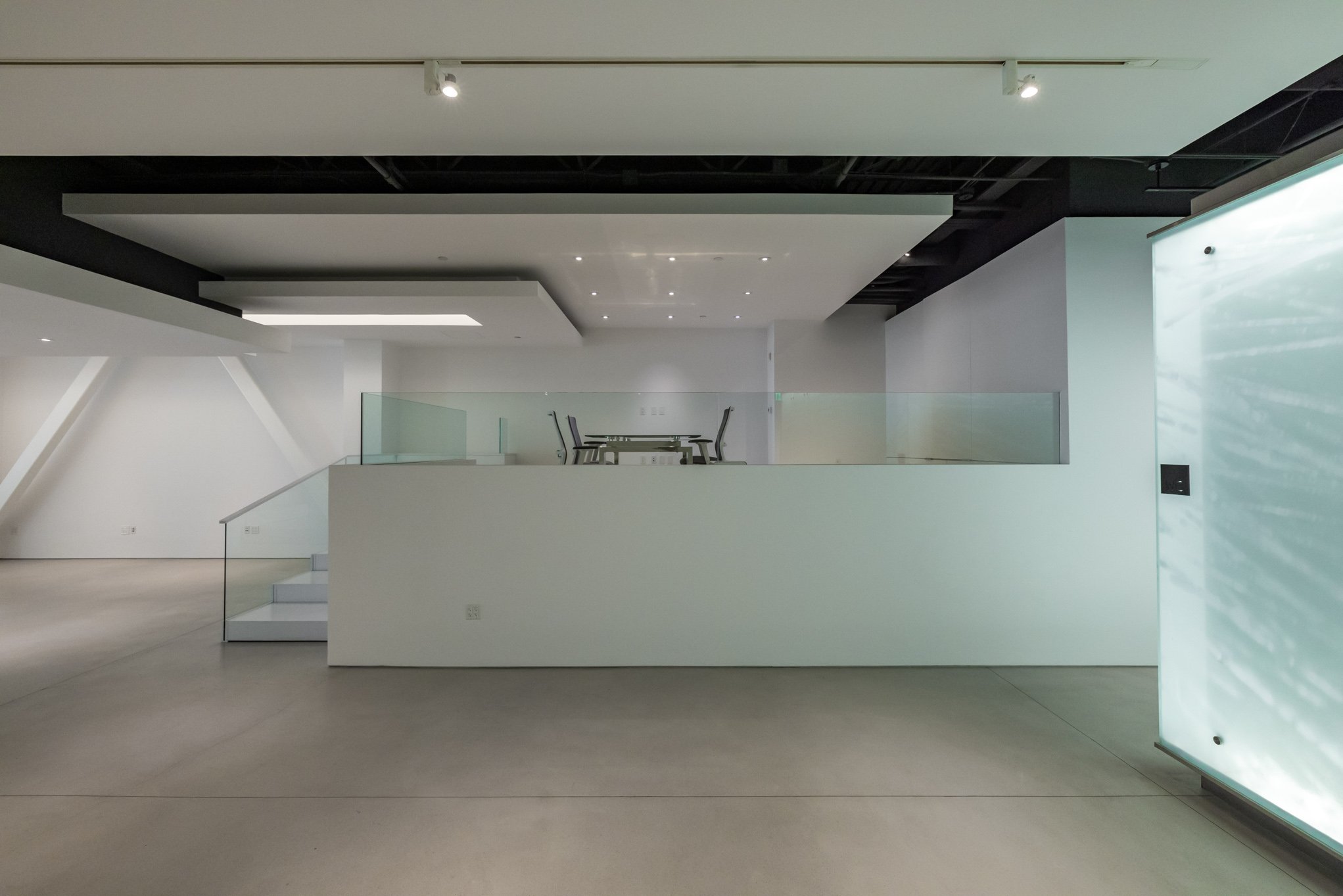 Interior view of a modern, minimalist space with white walls, a small staircase, black chairs and a glass railing.