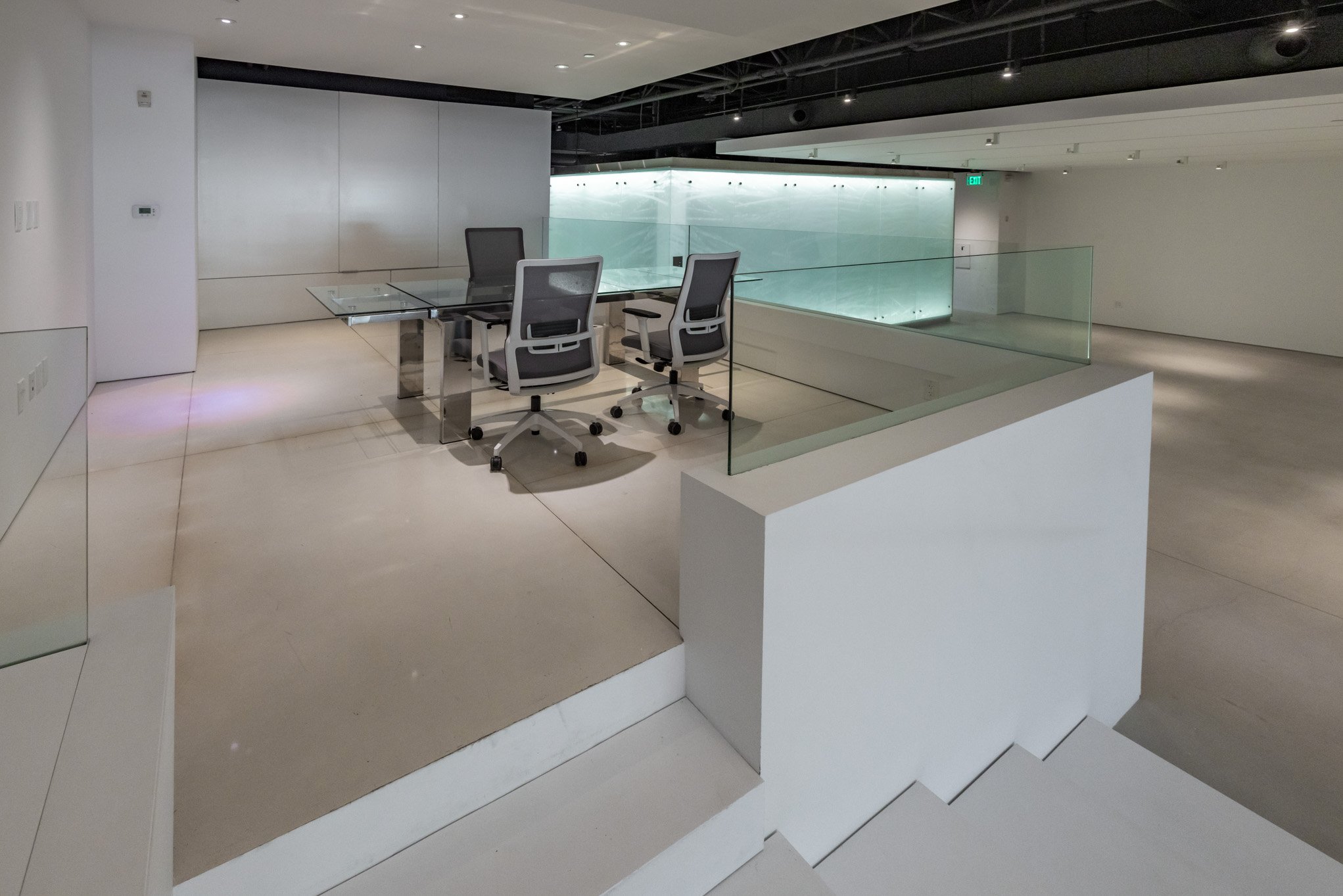 Empty modern office meeting room with glass table and four office chairs, overlooking a stairwell with glass railing in a minimalist building interior.