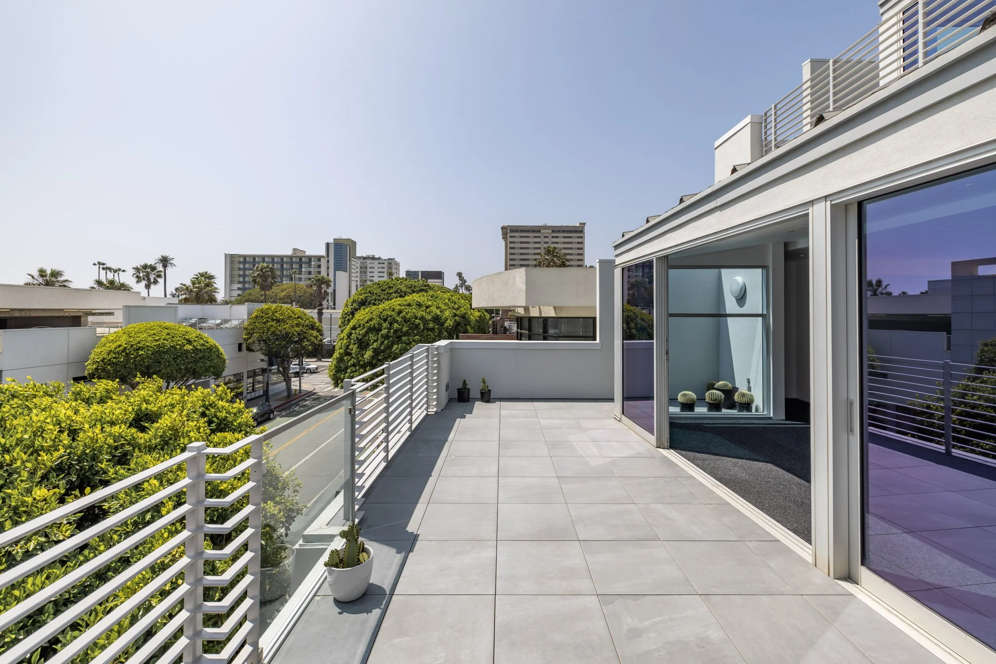 Empty balcony with white railing and potted plants, overlooking trees and city buildings in the distance, under a clear blue sky.