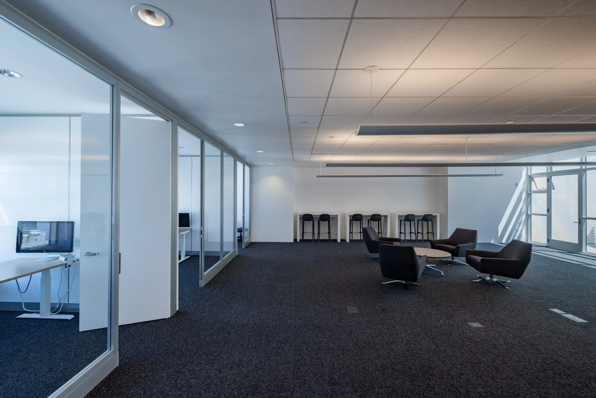 Empty modern office lounge area with black chairs and a table, glass-walled small office rooms on the left, and high stools at the back against a white wall, with sunlight coming through windows.