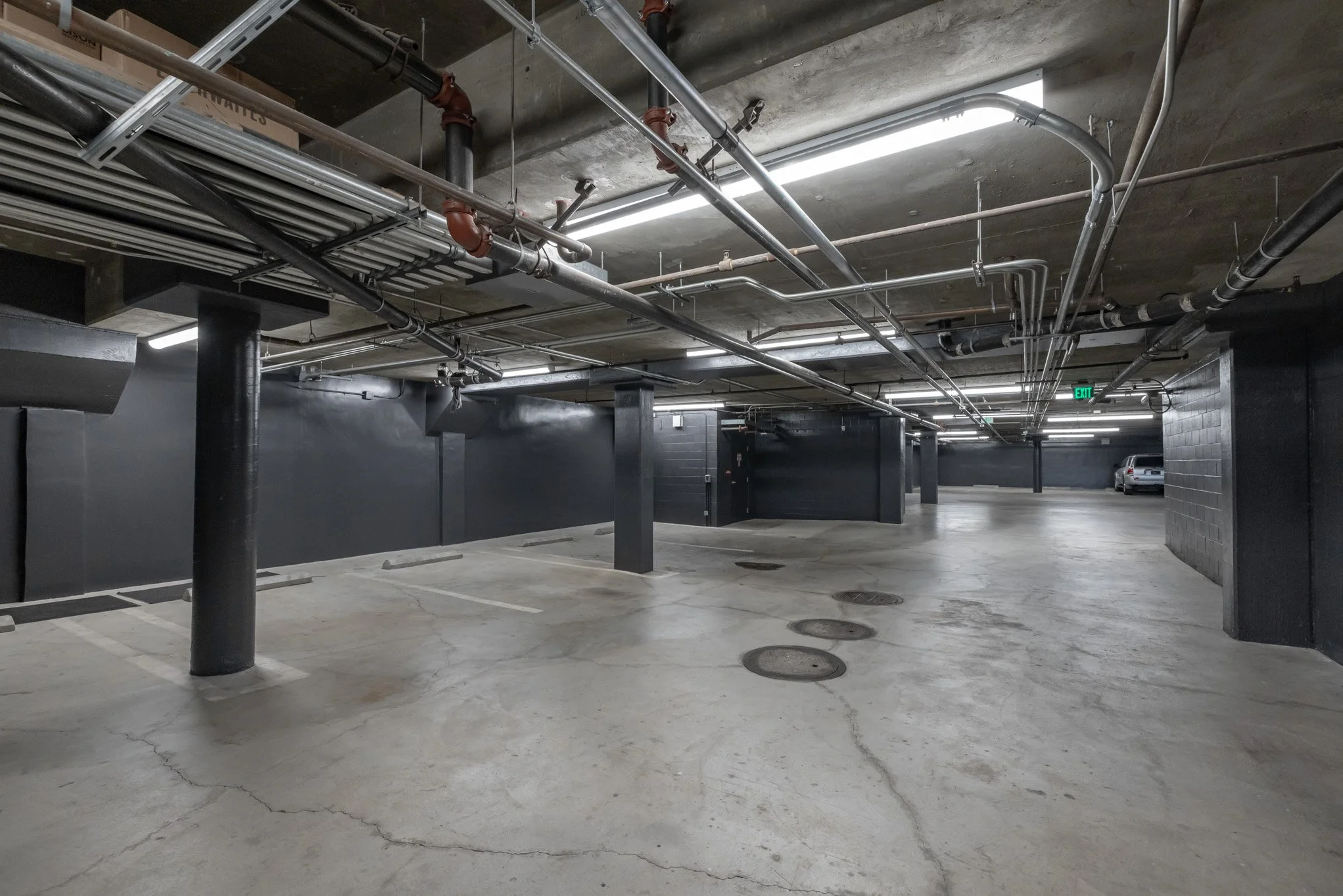 Underground parking garage with empty parking spaces, black painted walls and ceiling, exposed pipes, and a parked vehicle in the background.