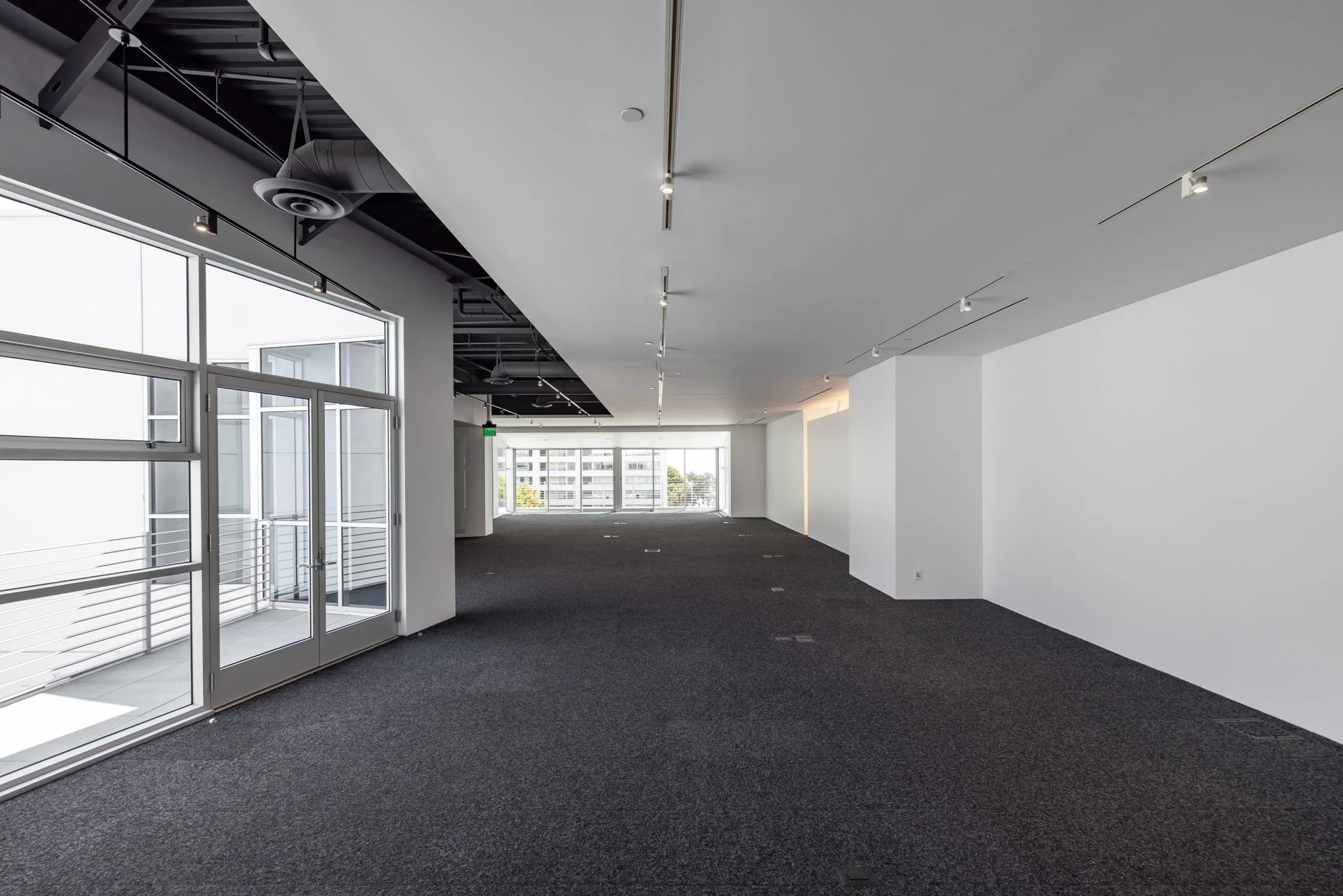 Empty office space with large windows, gray carpet, white walls, and ceiling with lighting fixtures.