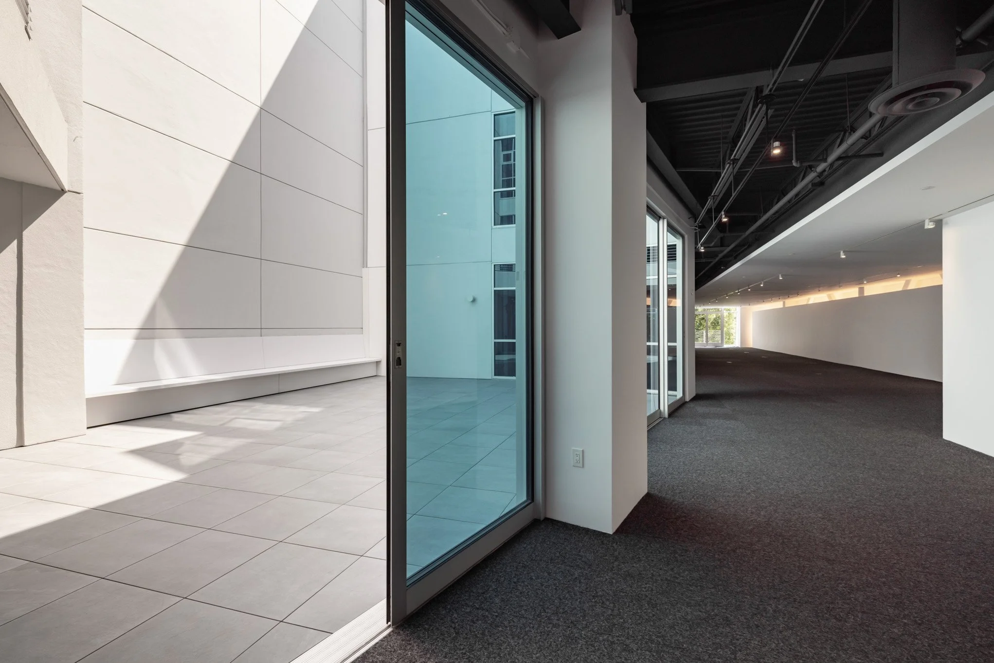 Empty modern room with large sliding glass doors opening to an outdoor patio area, featuring white walls, tiled floor, and a dark ceiling with ceiling-mounted fixtures.