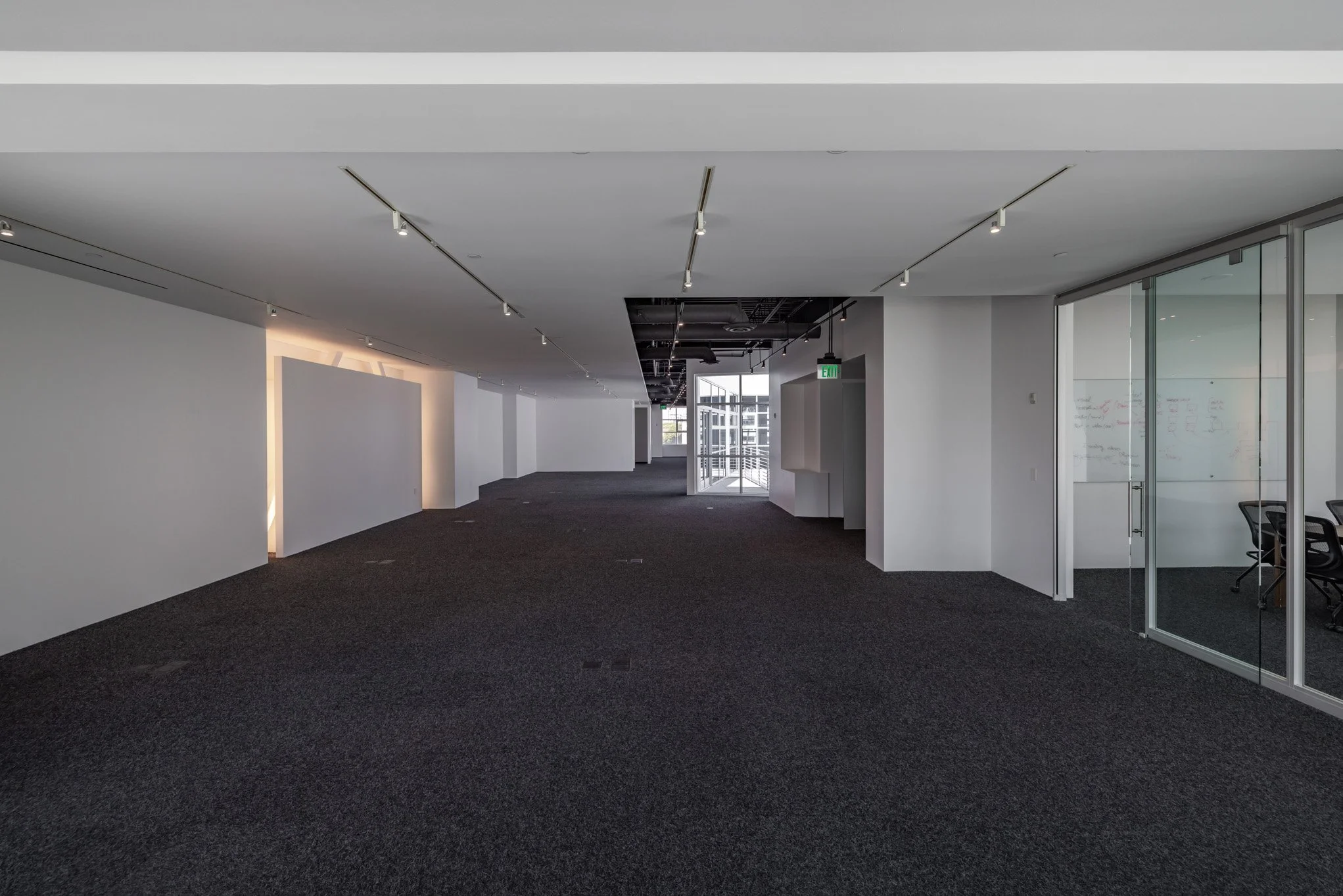 Empty office space with white walls, dark carpet, and glass walls on the right, with visible chairs inside, and an exit door in the background.