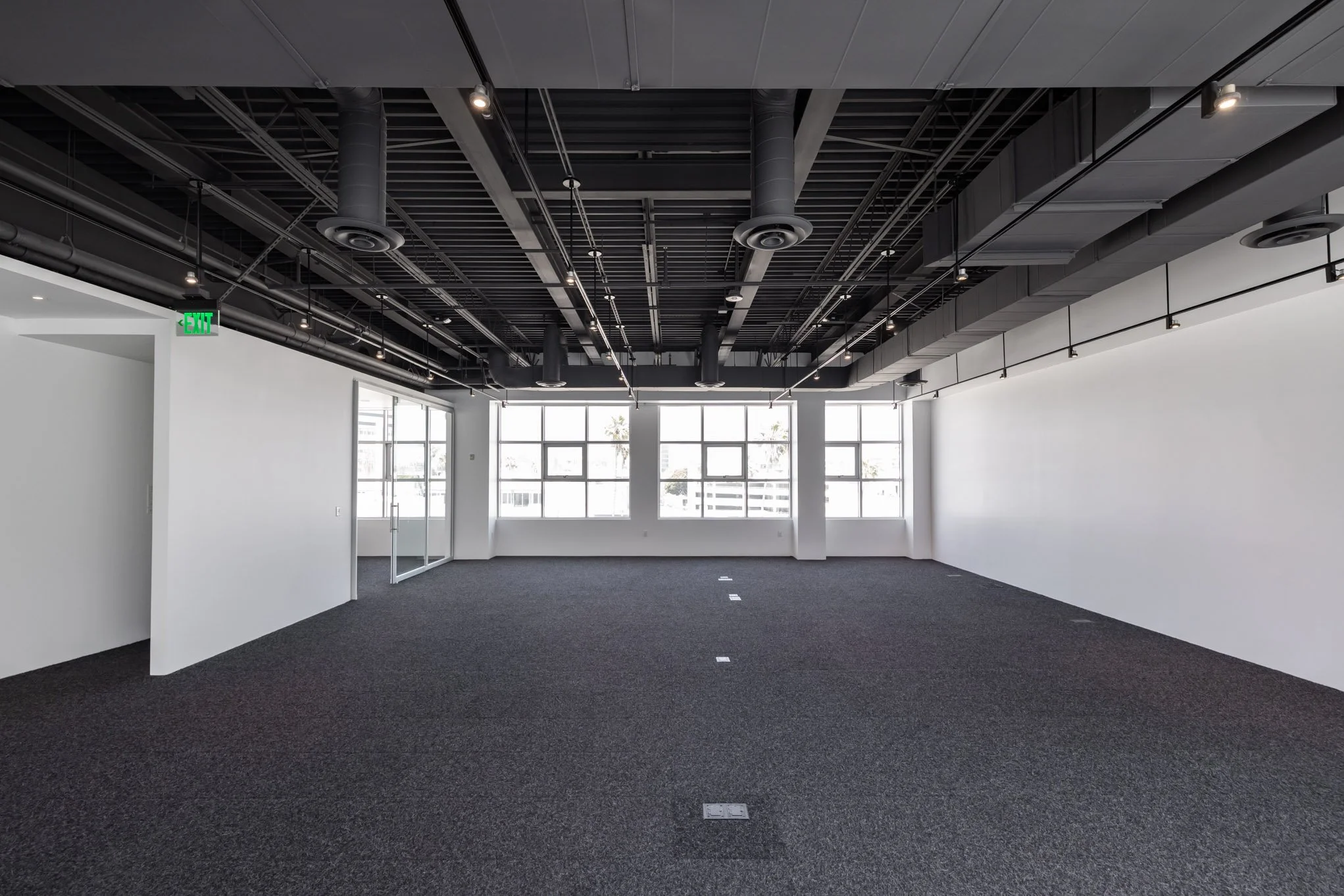 Empty office space with large windows, black ceiling with visible ducts and pipes, gray carpet flooring, and white walls.