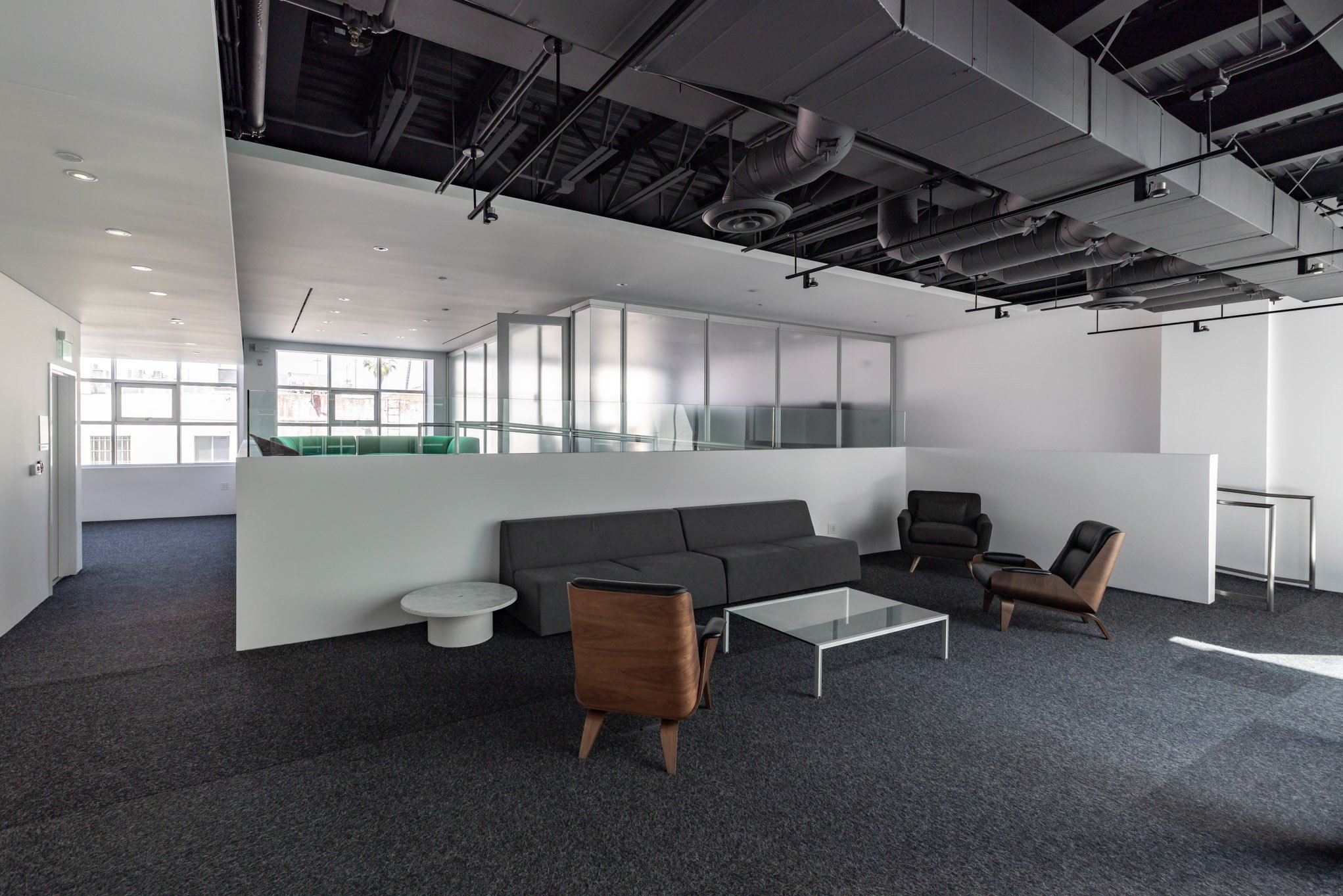 Modern office lounge with black, brown, and dark gray seating, a glass coffee table, carpeted floor, and large windows with natural light