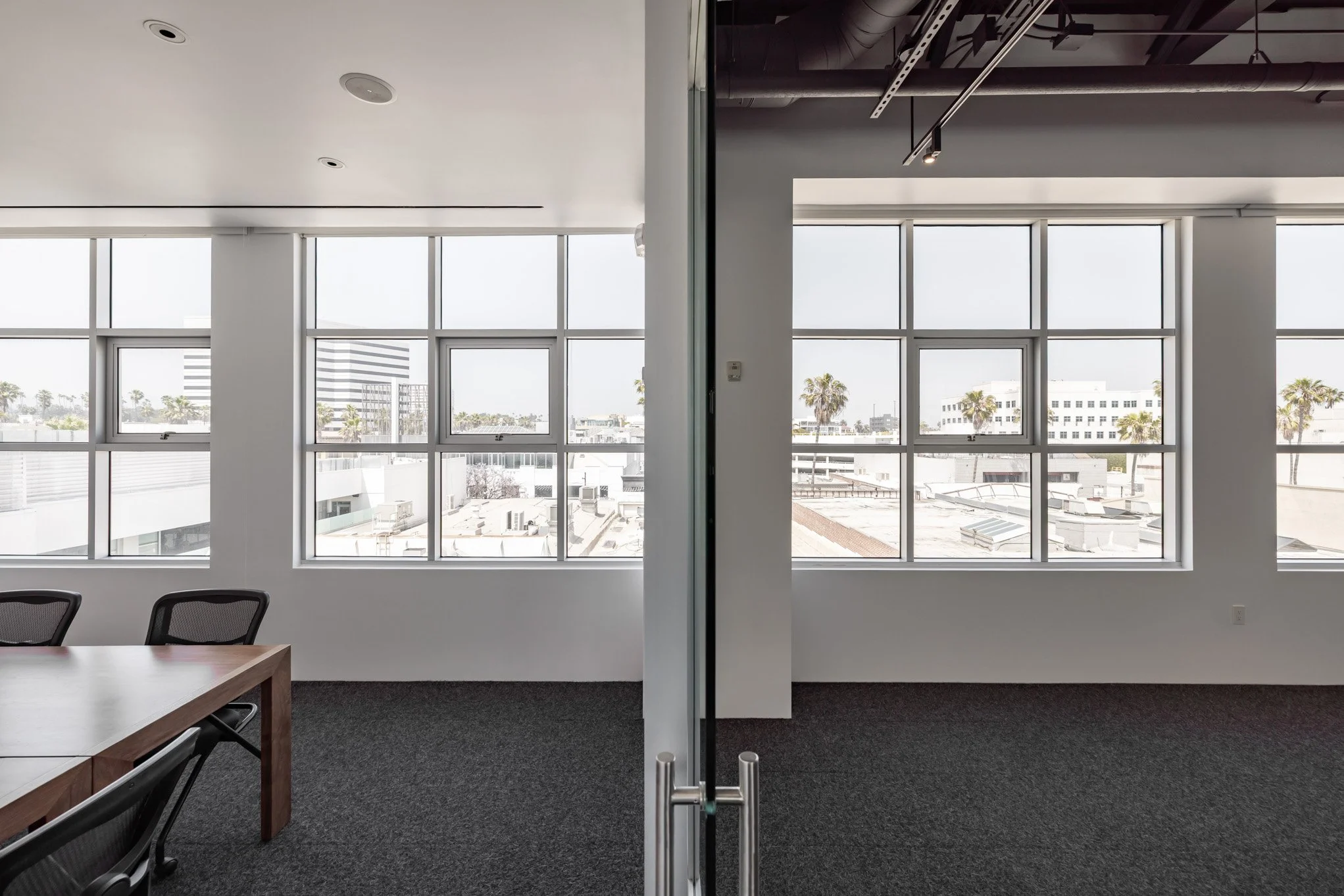 Split view of an office conference room with large windows, white walls, and modern ceiling, featuring a wooden table and black office chairs near the left side.