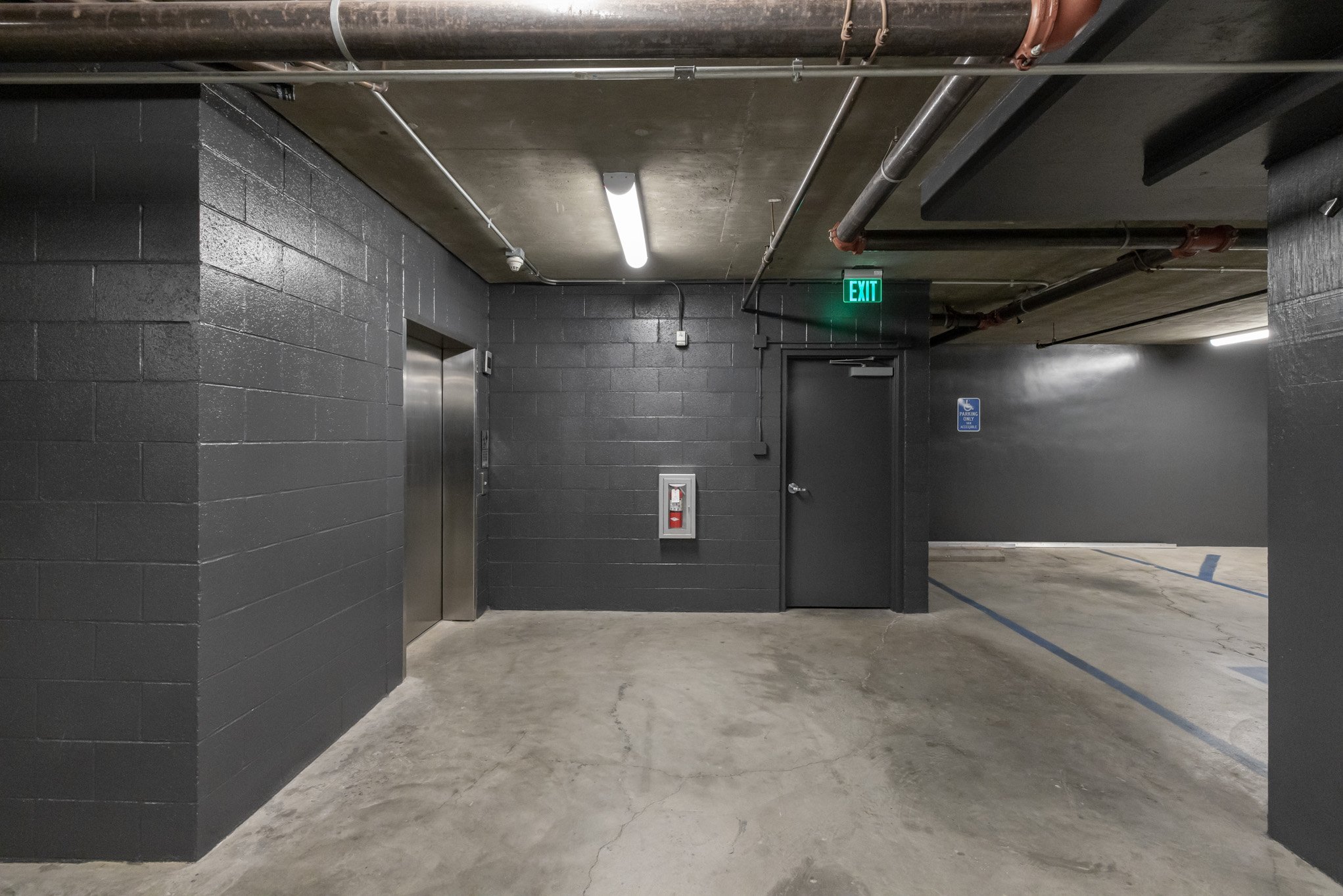 Underground parking garage with dark gray painted cinder block walls, concrete floor, an elevator door, an exit door with a green exit sign, and parking spots with blue lines.