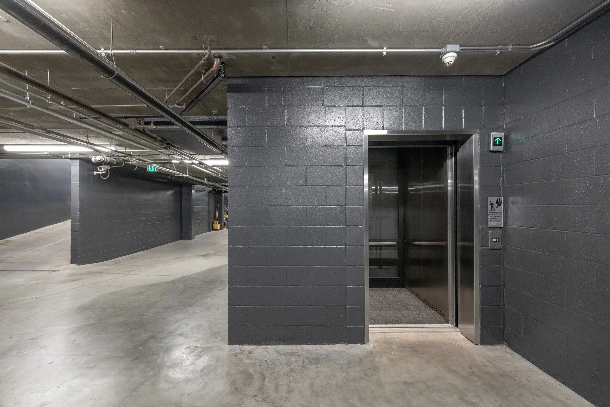 Elevator in a dark gray cinder block wall corridor with green up arrow indicator and safety sign.
