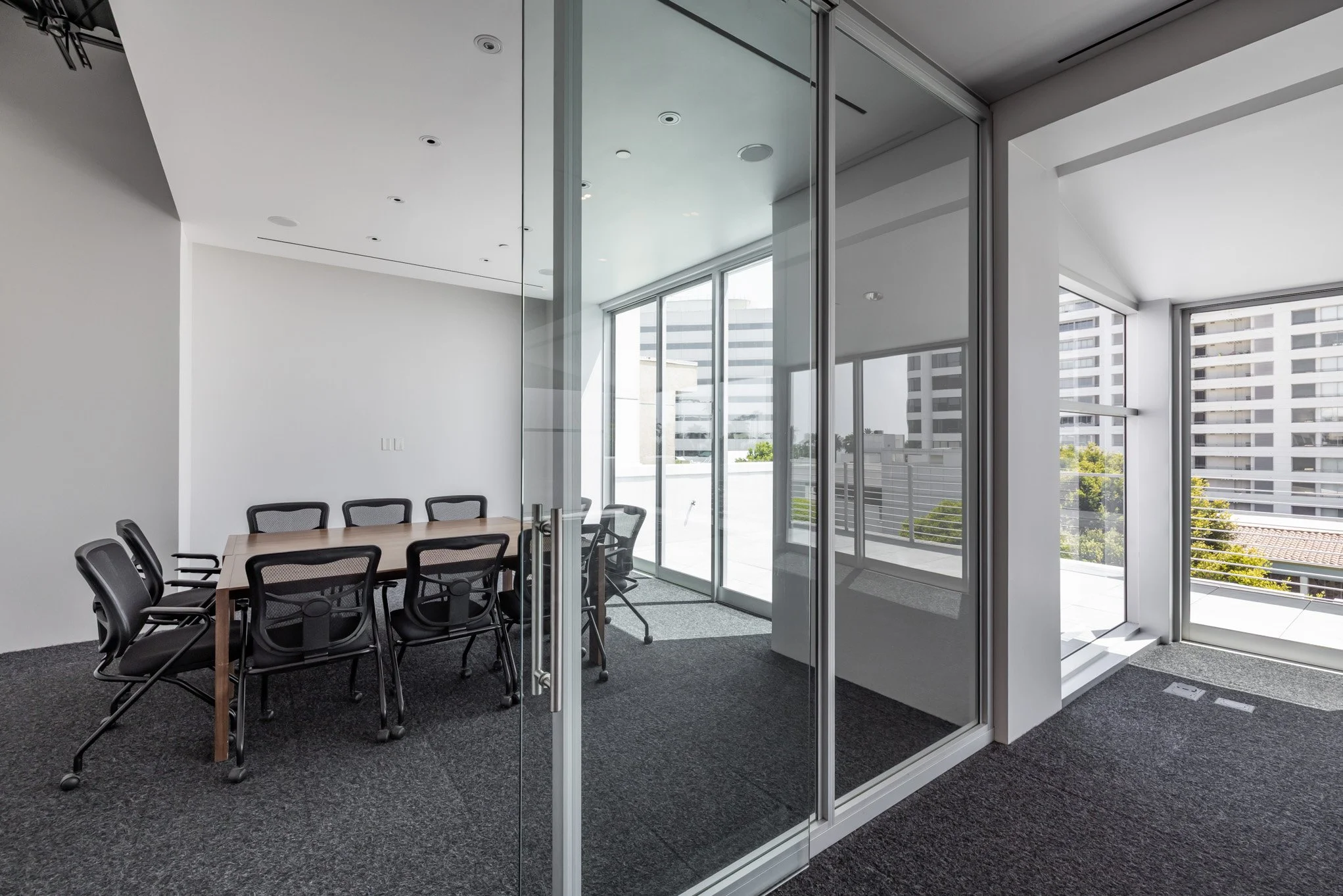 Empty modern conference room with glass walls, a wooden table, and black office chairs, with large windows overlooking city buildings.