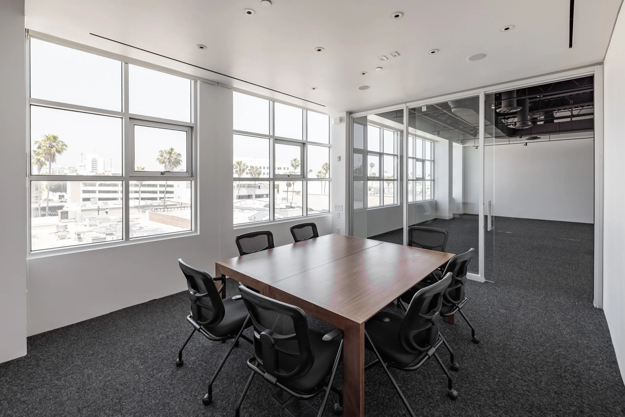 Empty modern conference room with a wooden table and six black office chairs, large windows showing a palm-lined cityscape, and a glass wall leading to another room.