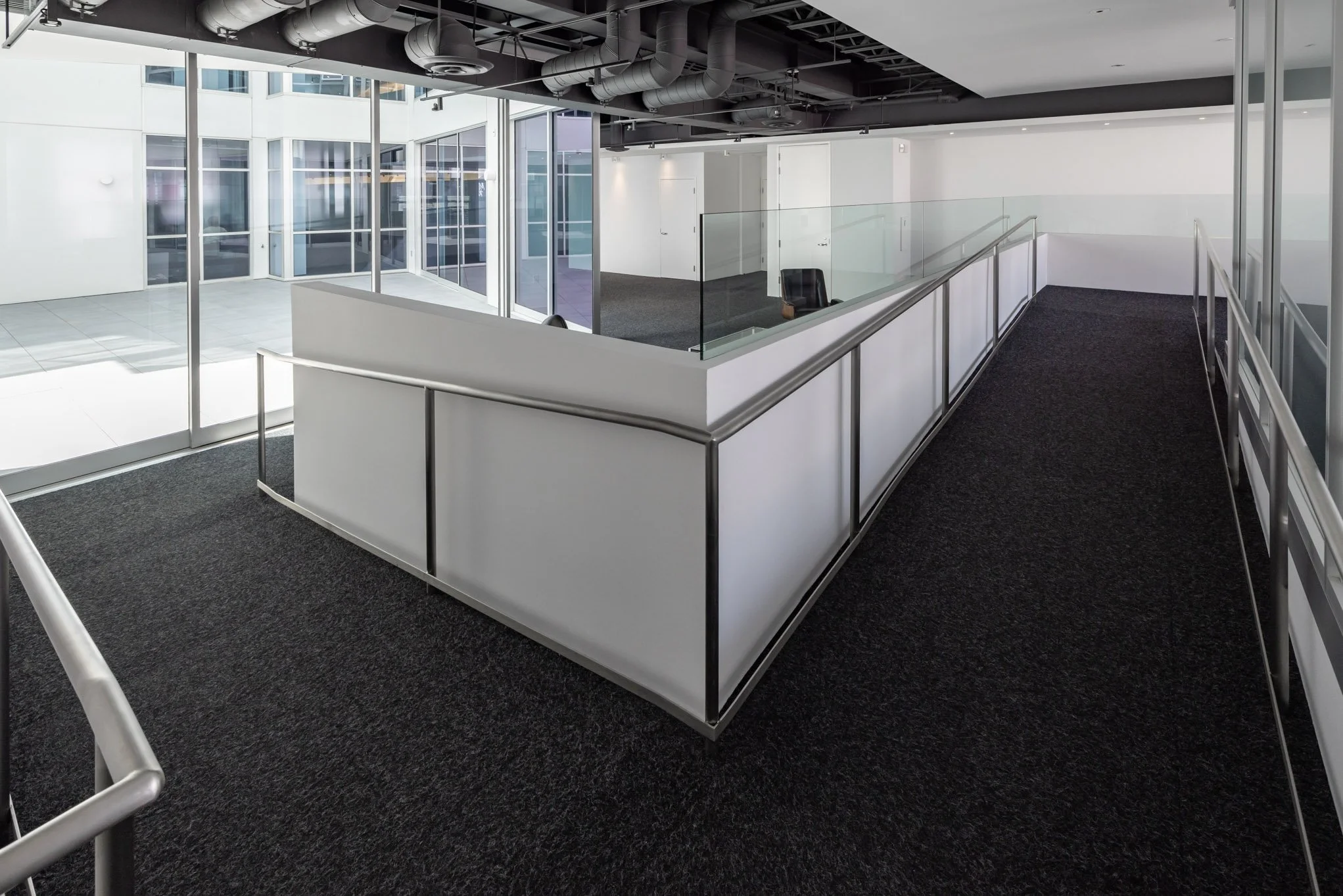 Empty modern office space with carpeted walkways, glass railing, and large windows letting in natural light.