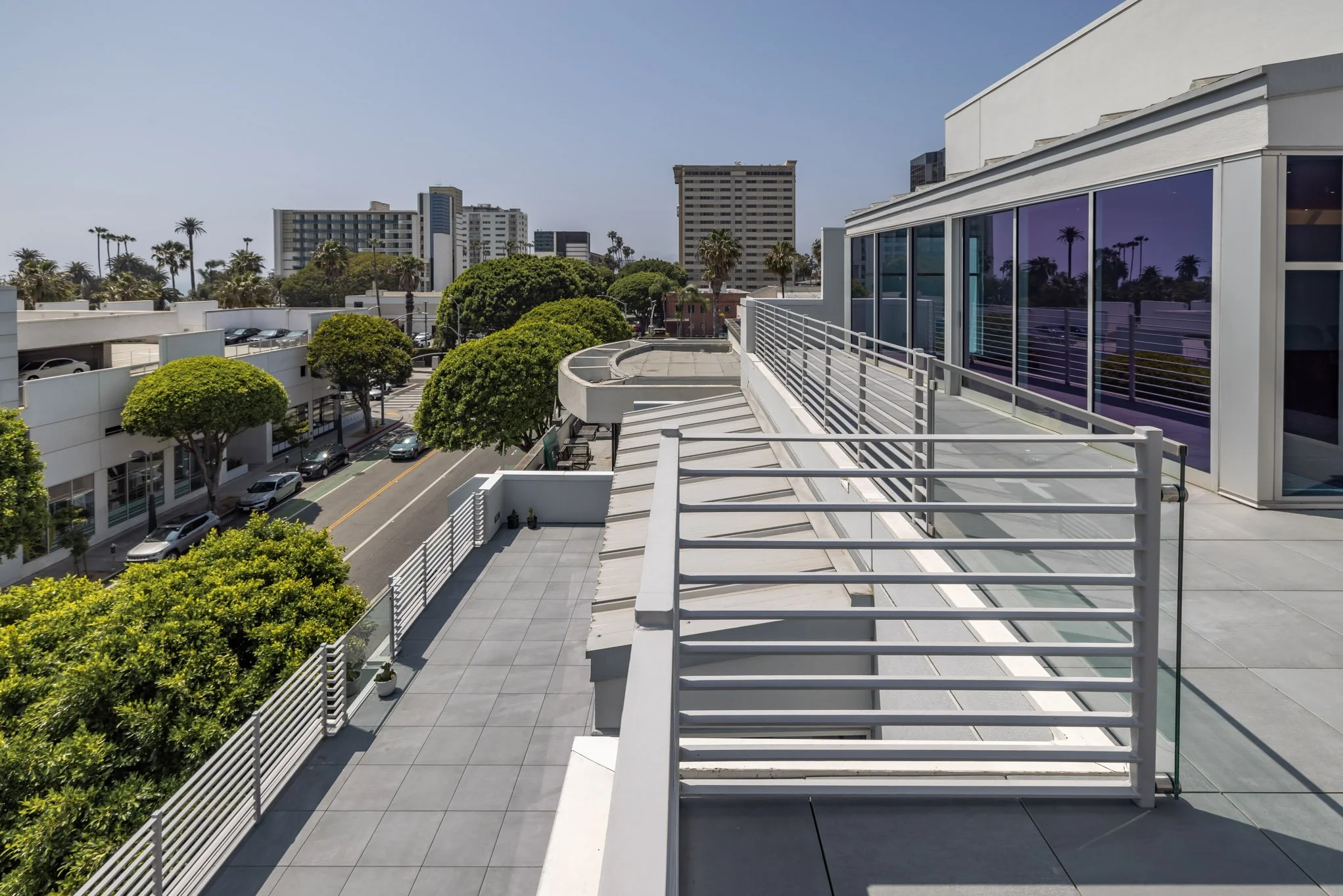 View from a rooftop terrace showing modern buildings, palm trees, parked cars, and a clear sky.
