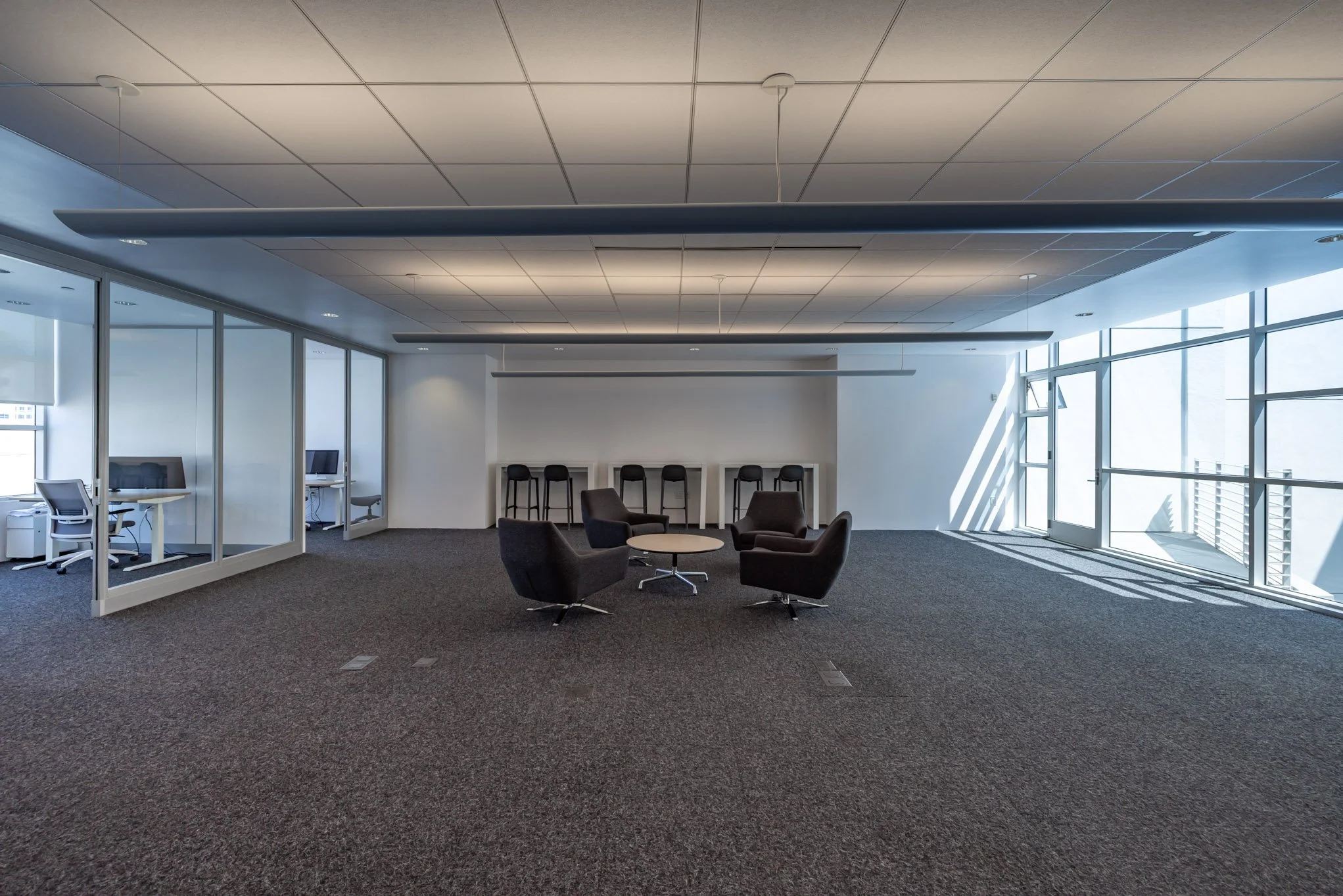 Empty modern office lounge area with four black chairs and a small round table in the center, multiple workstations with computers on the left, large windows on the right allowing natural light, and a white wall with bar stools at the back.