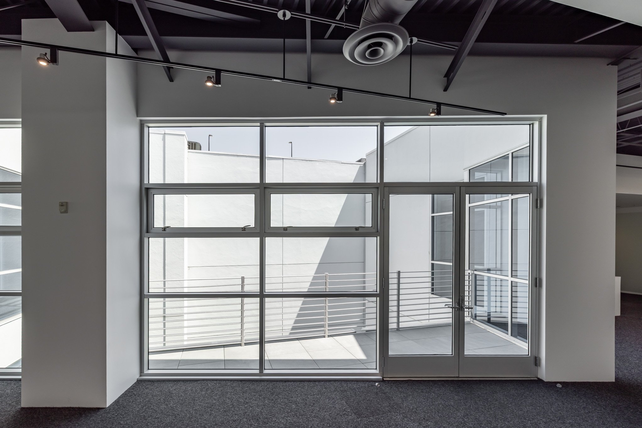 Modern interior space with large glass doors and windows, metal balcony railing, gray carpet, and ceiling with black beams and circular HVAC vent.