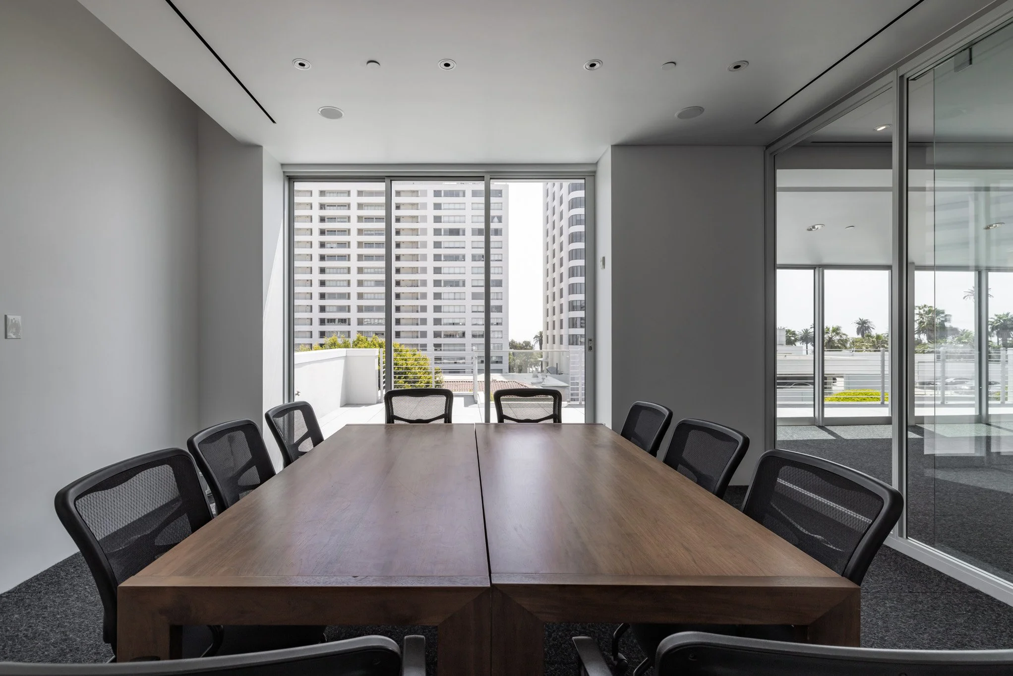 Empty conference room with a large wooden table, black mesh chairs, windows showing cityscape and balcony.