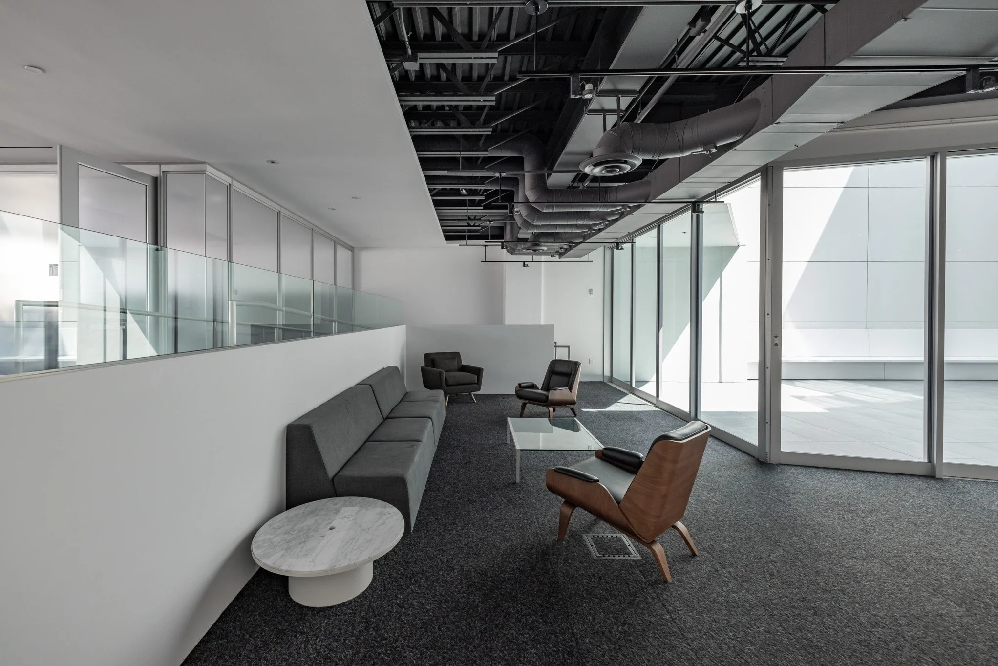 Modern office lounge area with gray sofa, black and brown chairs, glass coffee table, and large floor-to-ceiling windows with natural light.