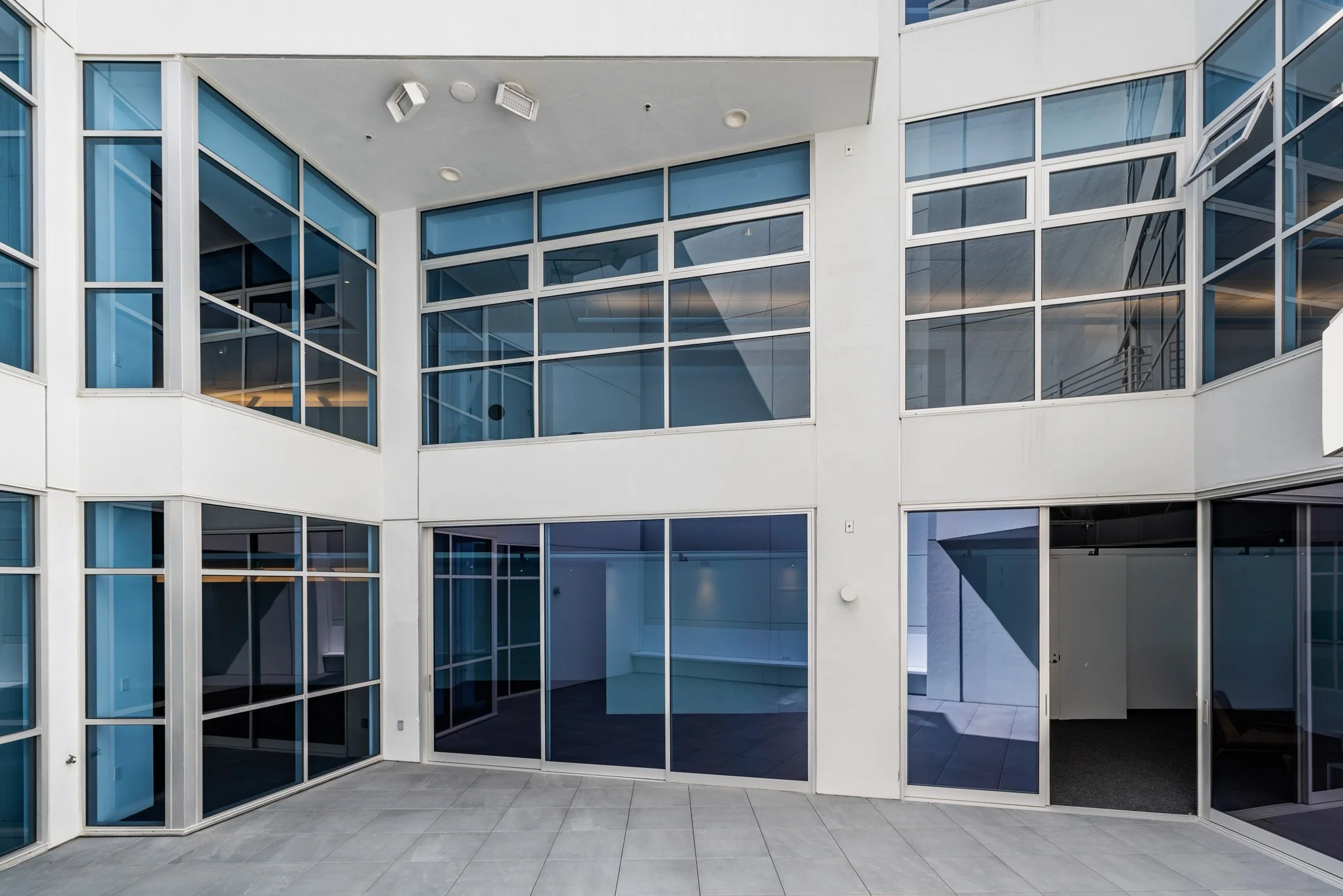 Modern interior courtyard with large glass windows and doors, white walls, tiled floor, and some ceiling lights.