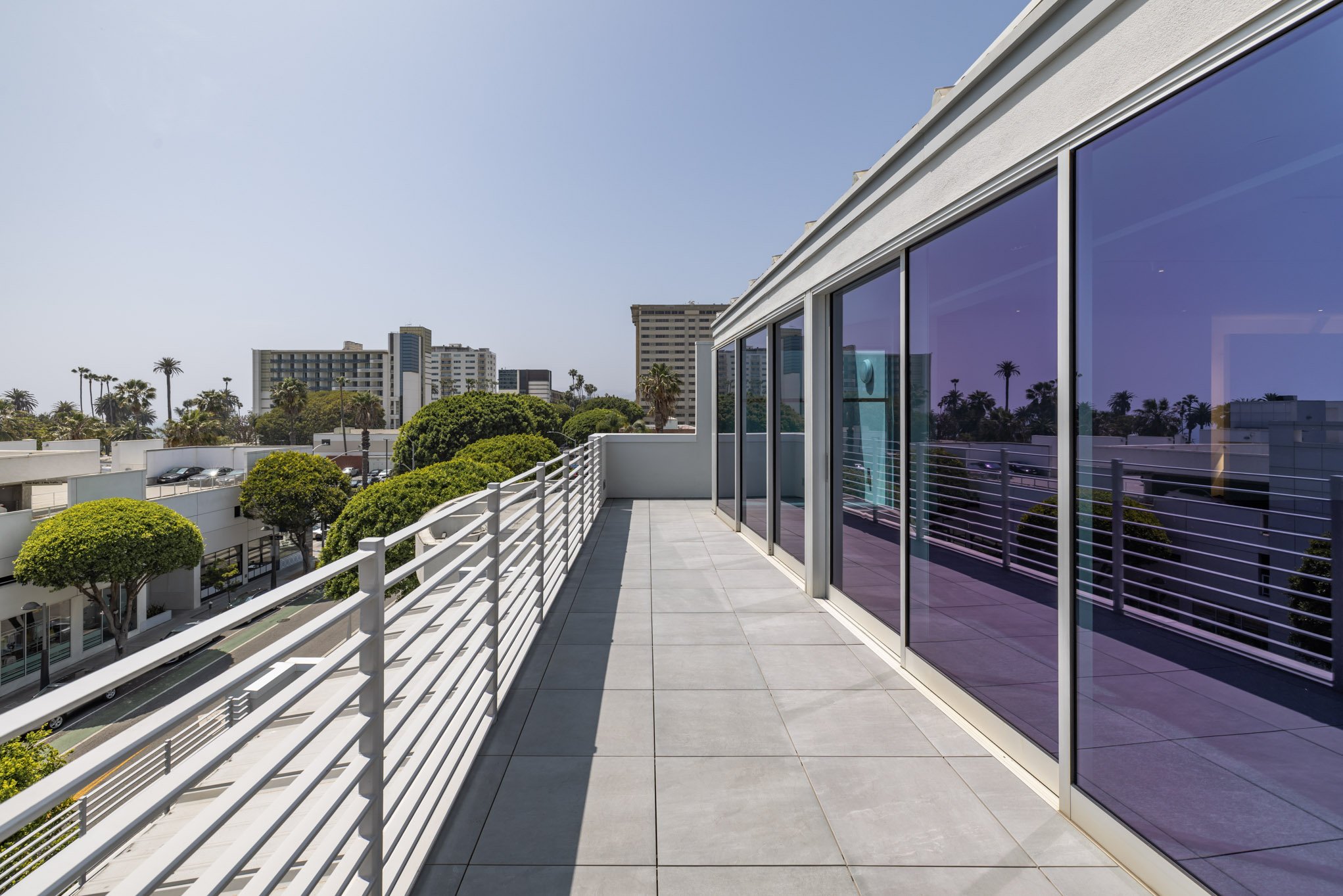 Empty balcony with glass sliding doors and city skyline in the background, featuring trees, buildings, and clear sky.