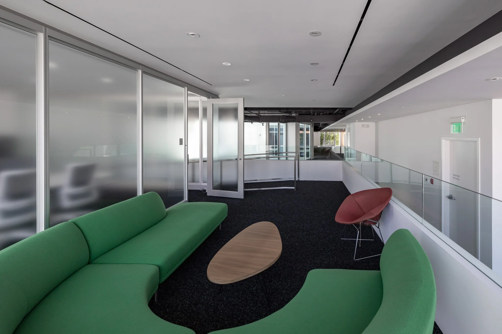 Modern office hallway with green curved sofa, red chair, and wooden coffee table, glass walls and open door leading to another room.