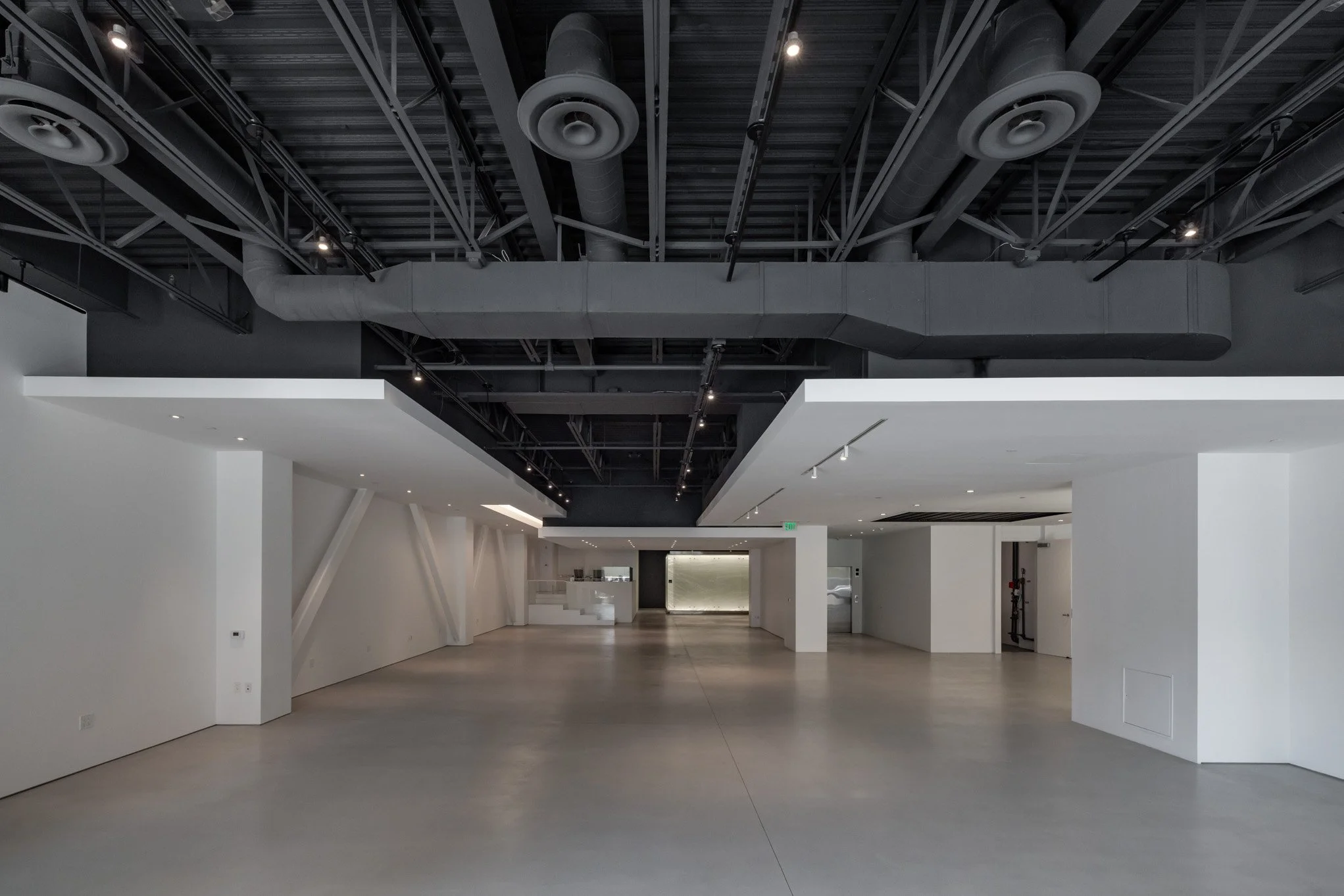 Empty modern indoor space with white walls and gray concrete floors, featuring an exposed ceiling with black painted ductwork and industrial lighting fixtures.