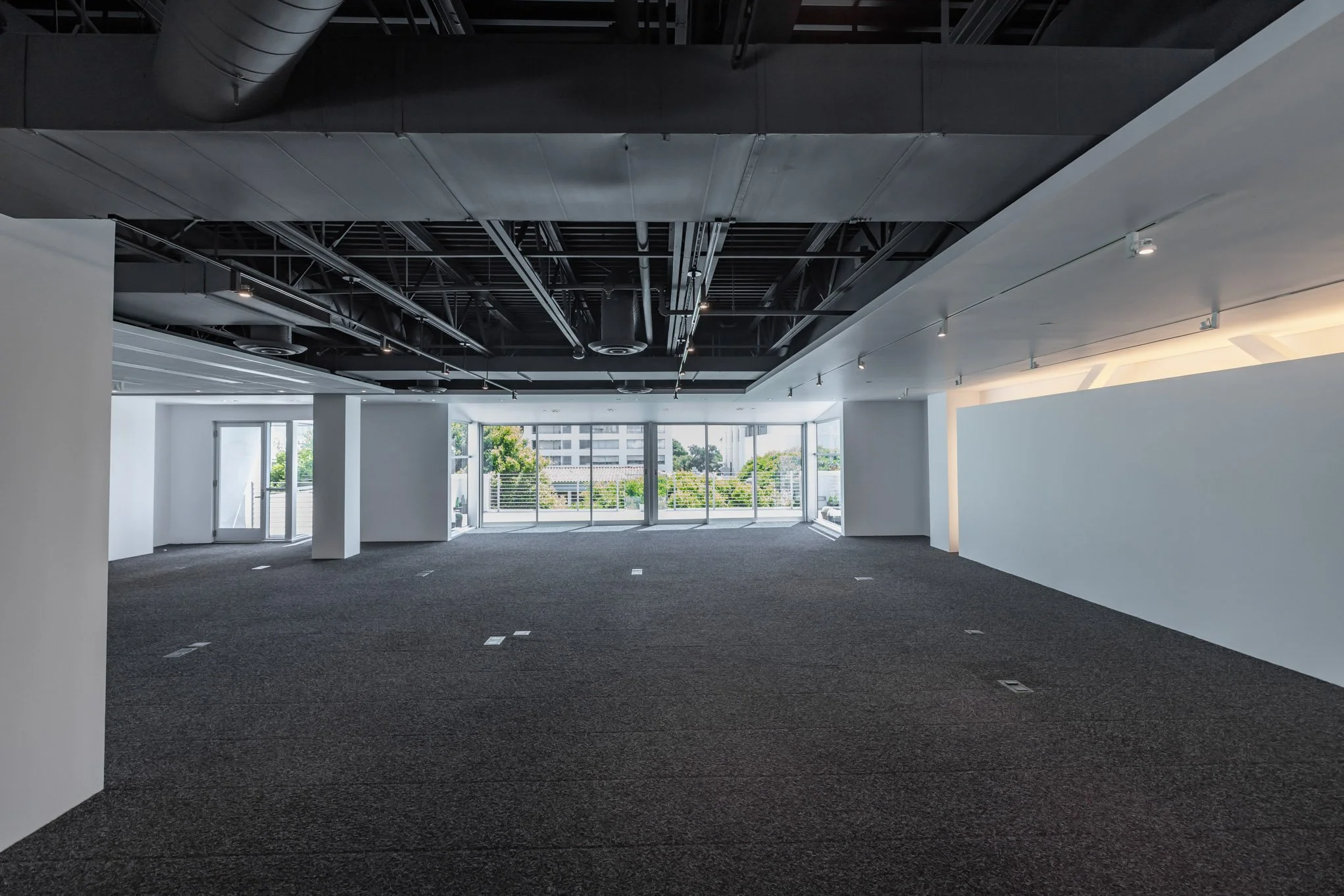 Empty commercial office space with large windows, gray carpet, white walls, and an exposed ceiling with visible ducts and piping.