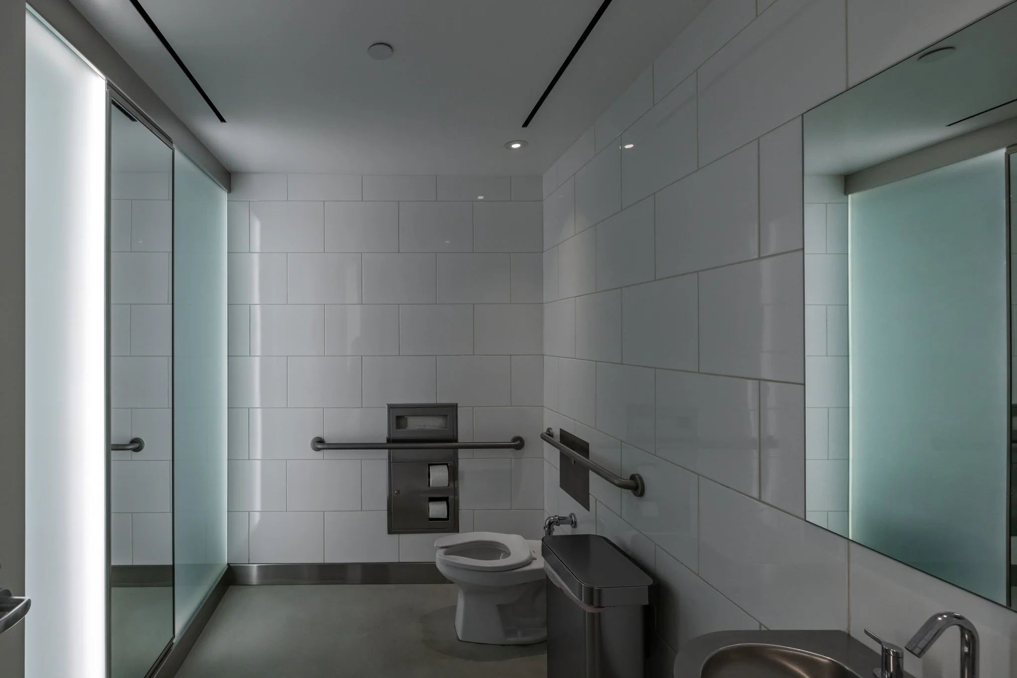 Accessible public bathroom with white tiled walls, a toilet, grab bars, a large mirror, and a frosted glass shower enclosure.