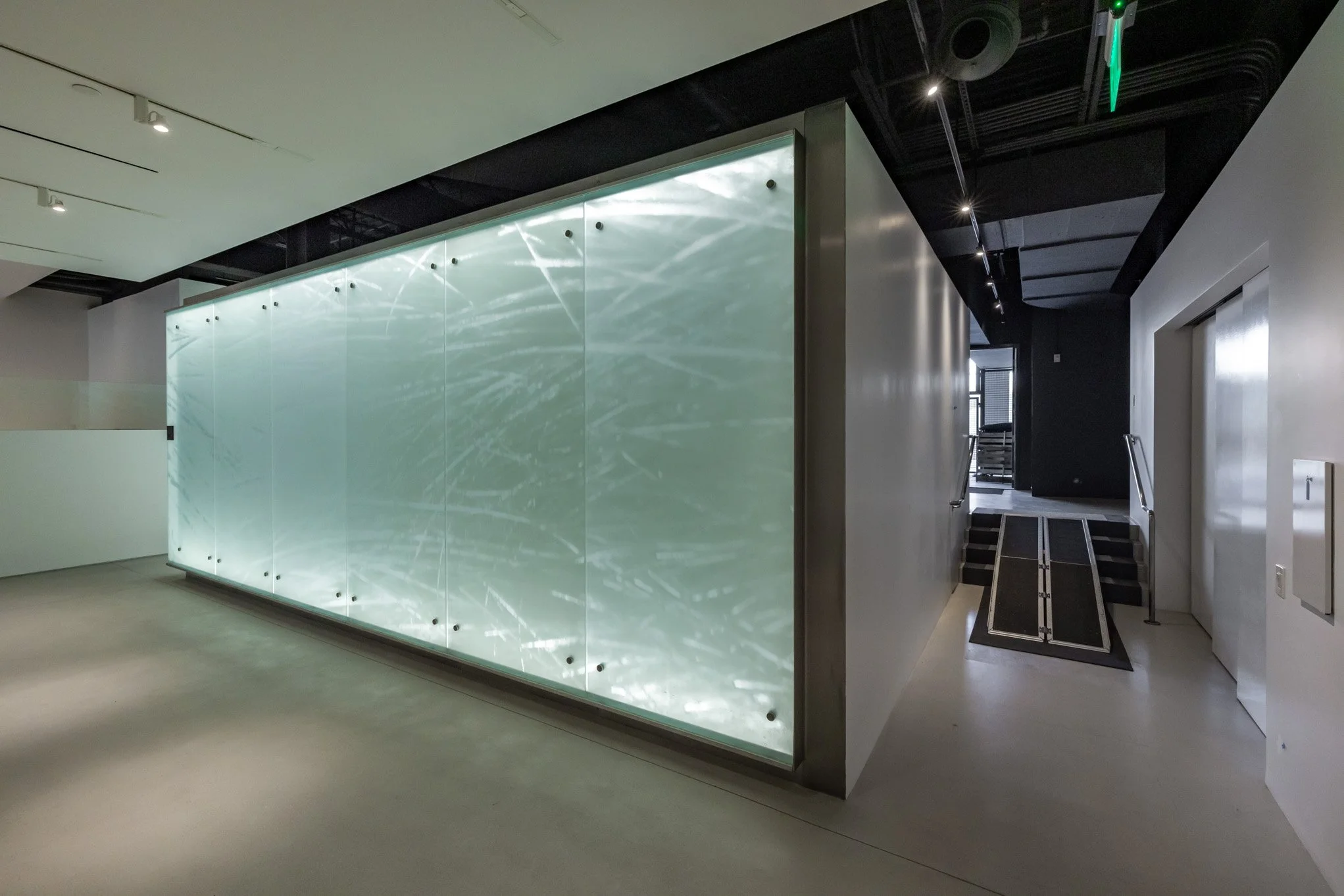 Modern interior with a large illuminated glass panel with water ripple pattern, black ceiling with track lighting, adjacent staircase with handrails, and white walls.