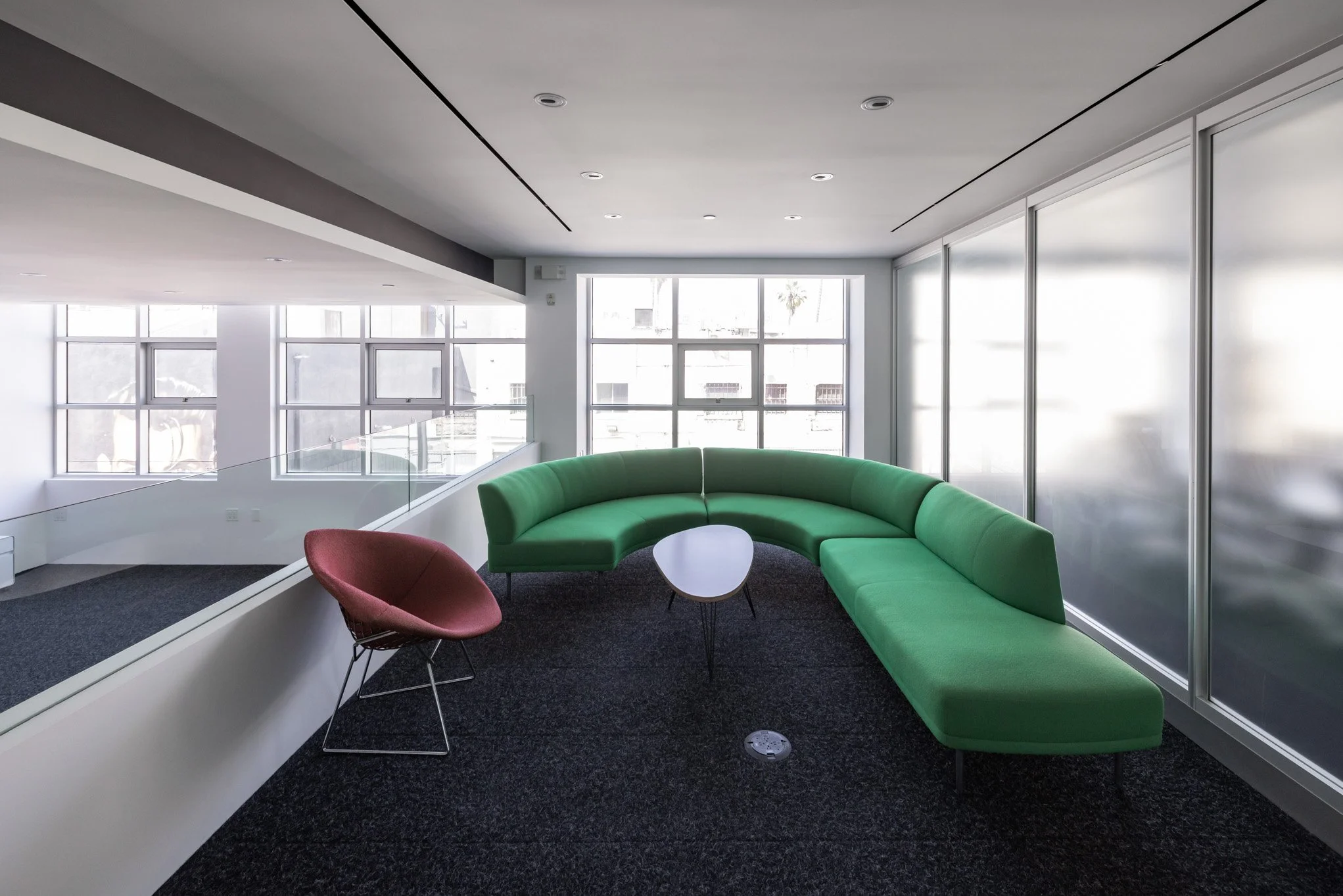 Modern office lounge with a curved green sofa, a pink accent chair, a small white coffee table, and large windows letting in natural light.
