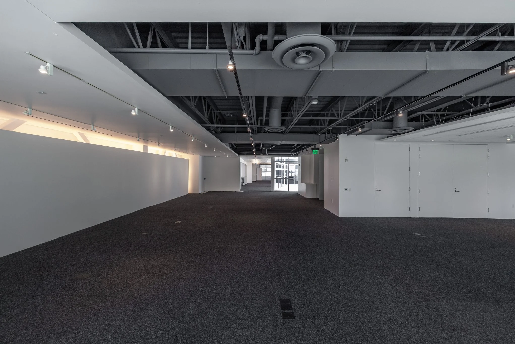 Empty modern office interior with white walls, black carpeted floor, exposed ceiling with ventilation ducts, and a glass door at the far end.