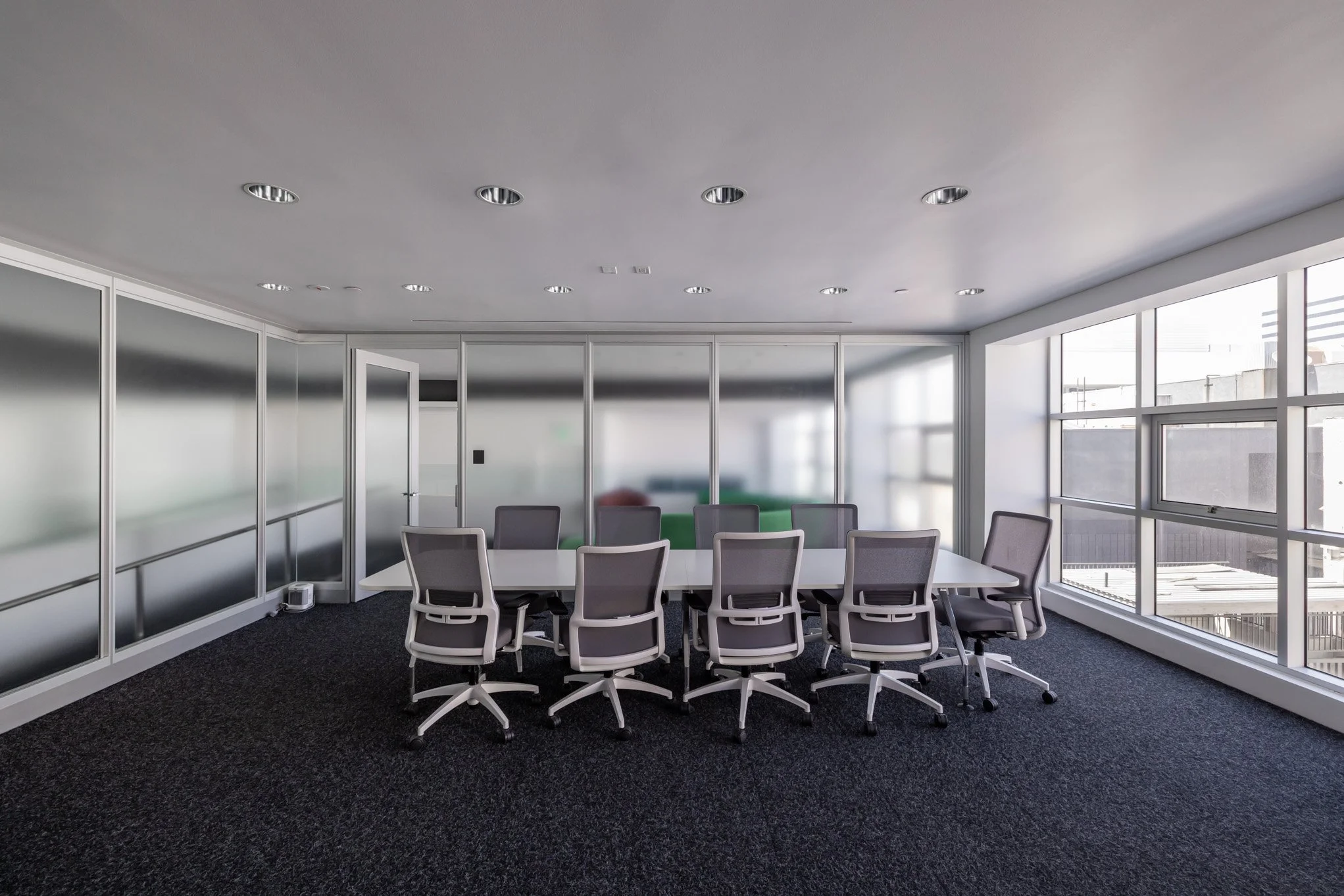 Modern conference room with a rectangular table and ten gray office chairs. Large windows on the right let in natural light. Frosted glass partitions on the far wall, with a white door slightly open on the left.